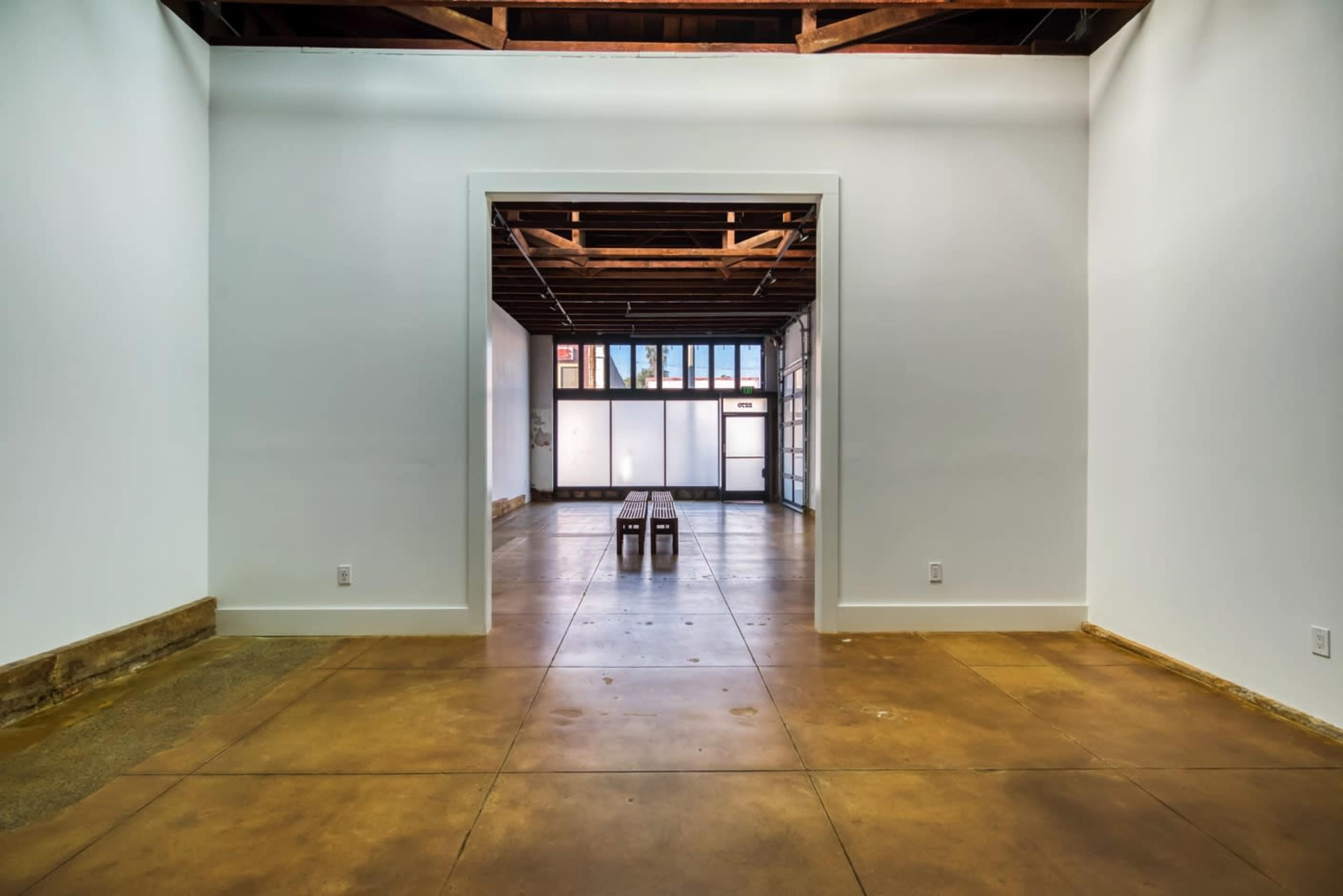 An interior space with a concrete floor and two wooden benches positioned centrally, leading to another room with large windows and exposed beams.