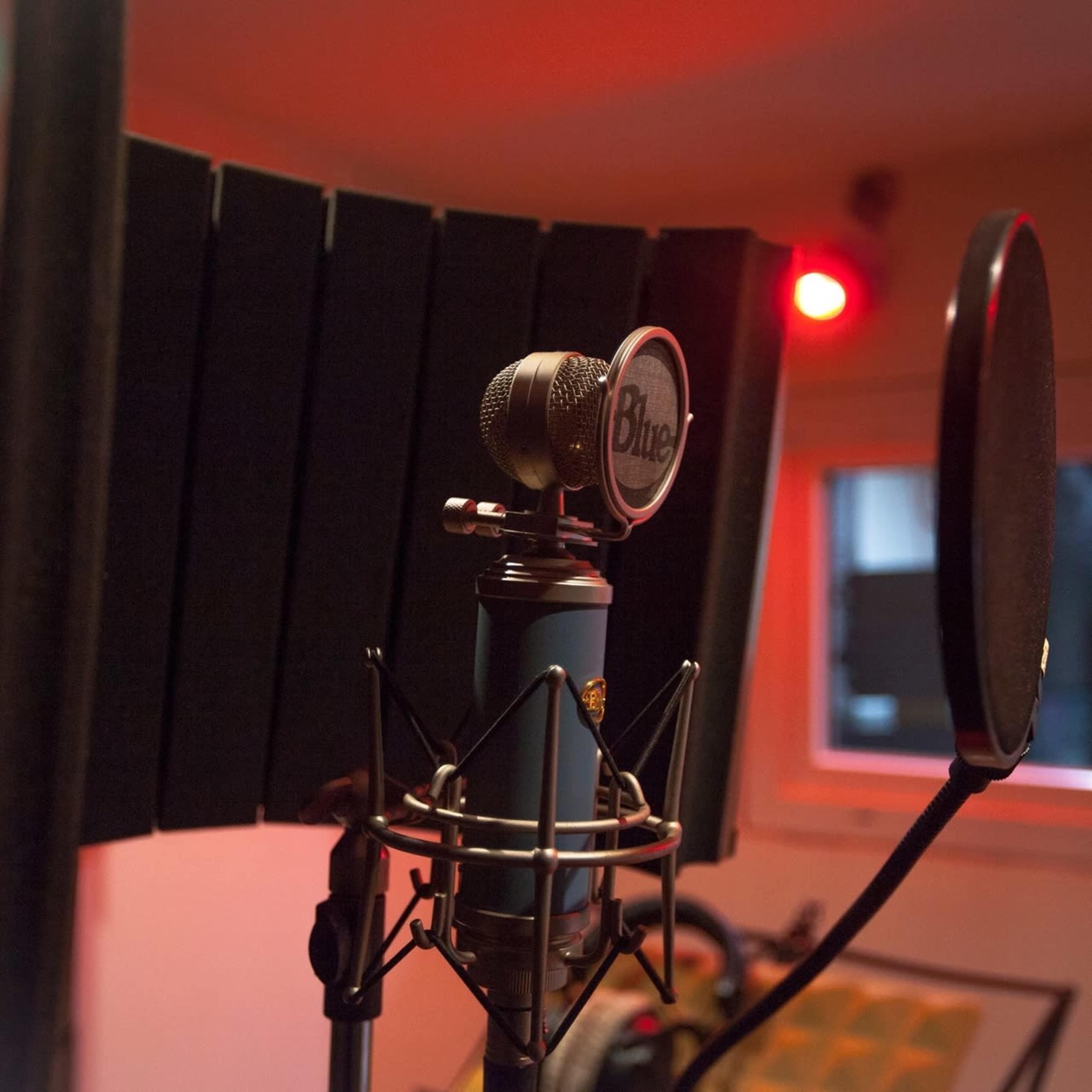 A microphone with a pop filter is set up in a recording studio, with soundproof panels and dim lighting in the background.
