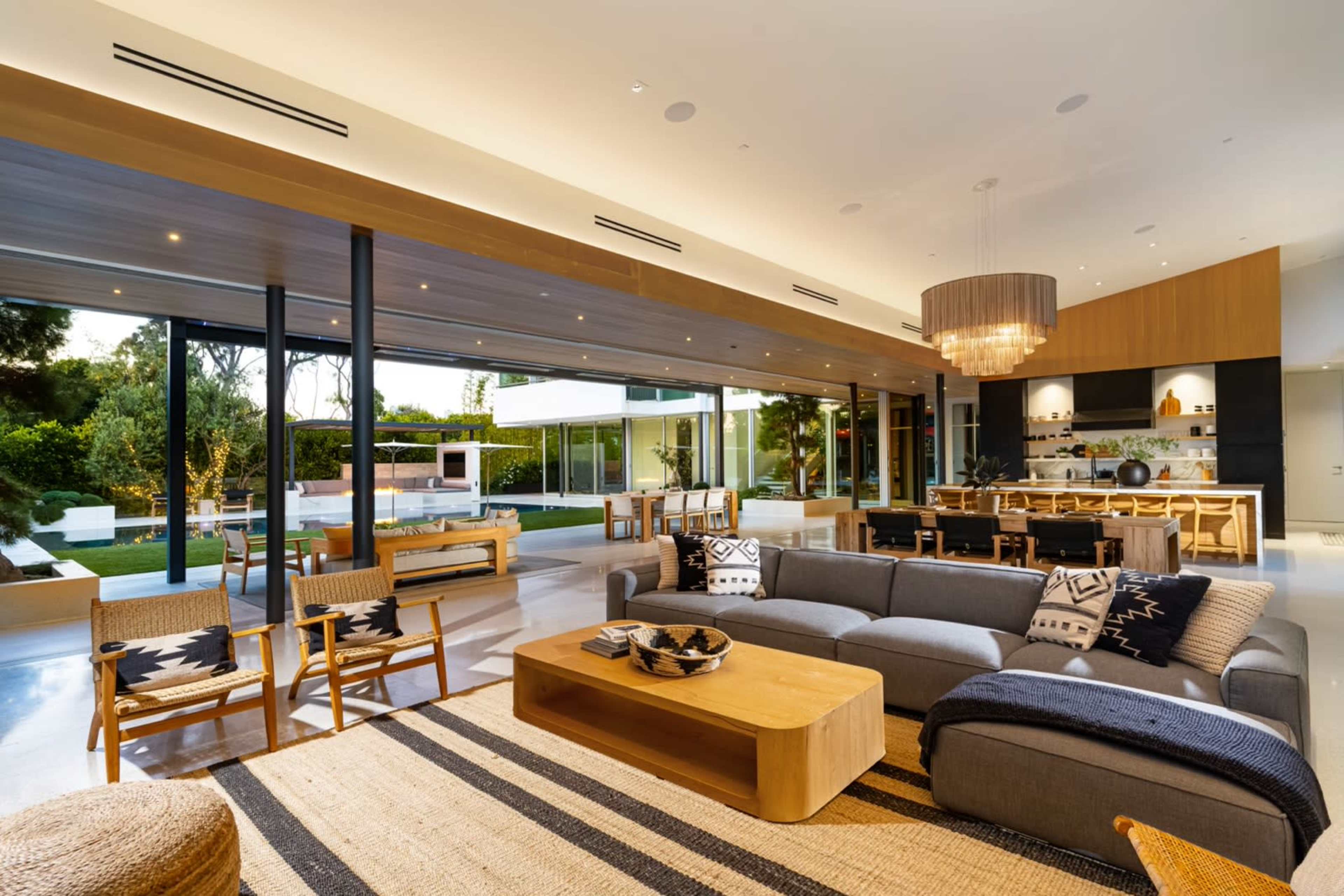 A modern open-concept living area with a large gray sofa, wooden dining table, and large glass windows overlooking a landscaped outdoor space.