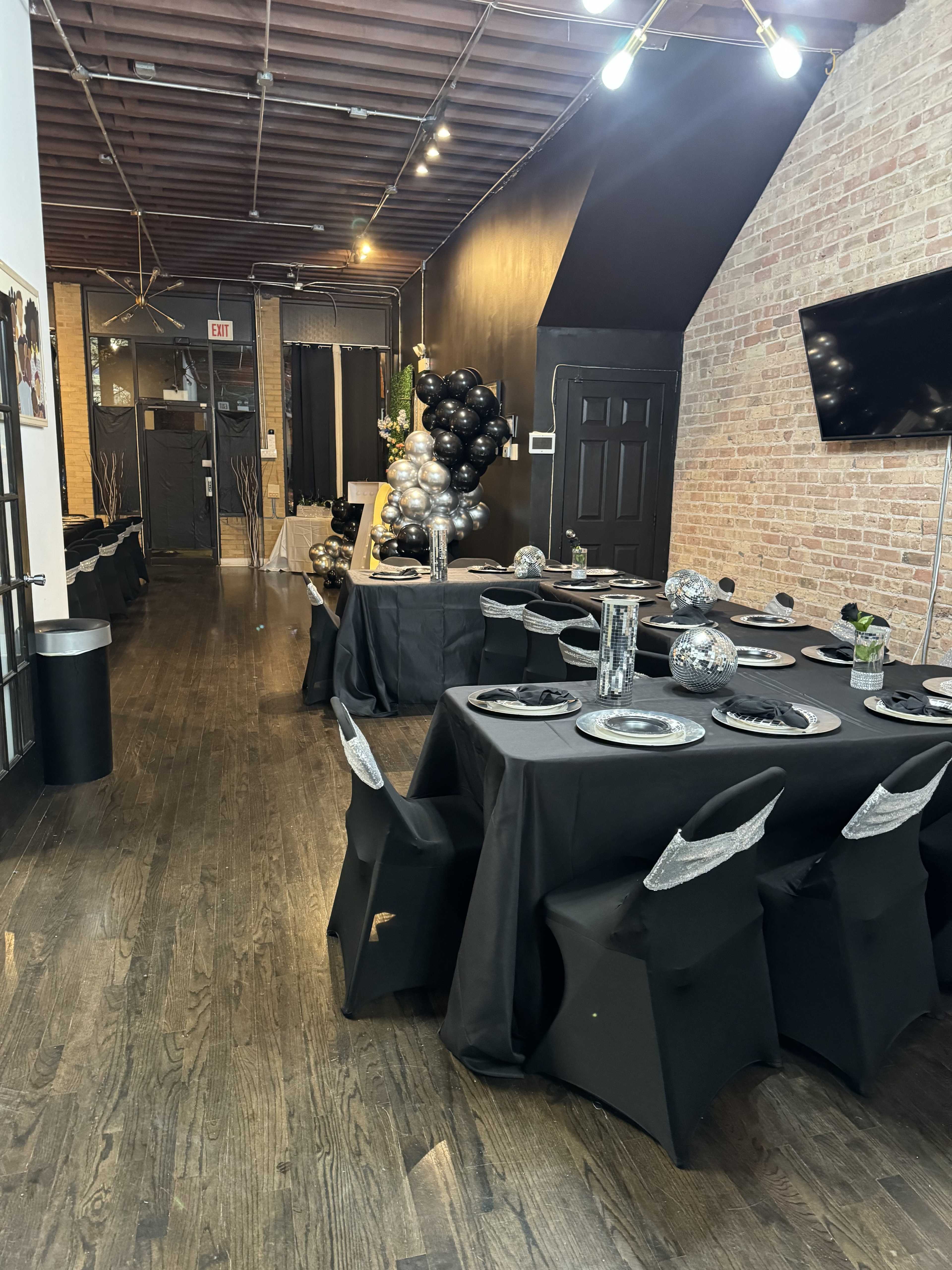 A dimly lit event space with black tables set for dining, decorated with silver and black balloons, and featuring a large screen on the wall.