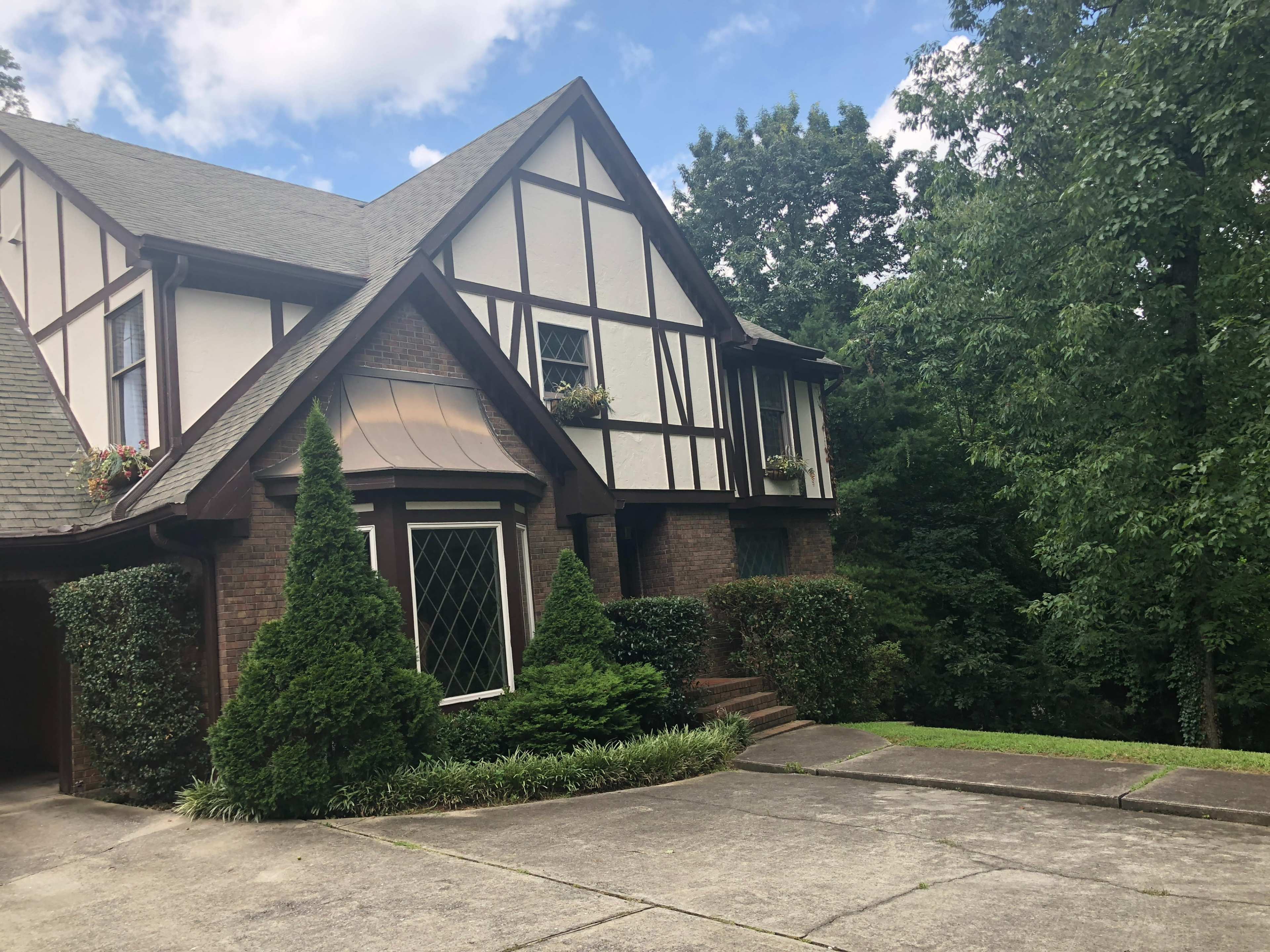 A two-story Tudor-style house with a brick and wood exterior, surrounded by greenery and featuring a paved driveway.