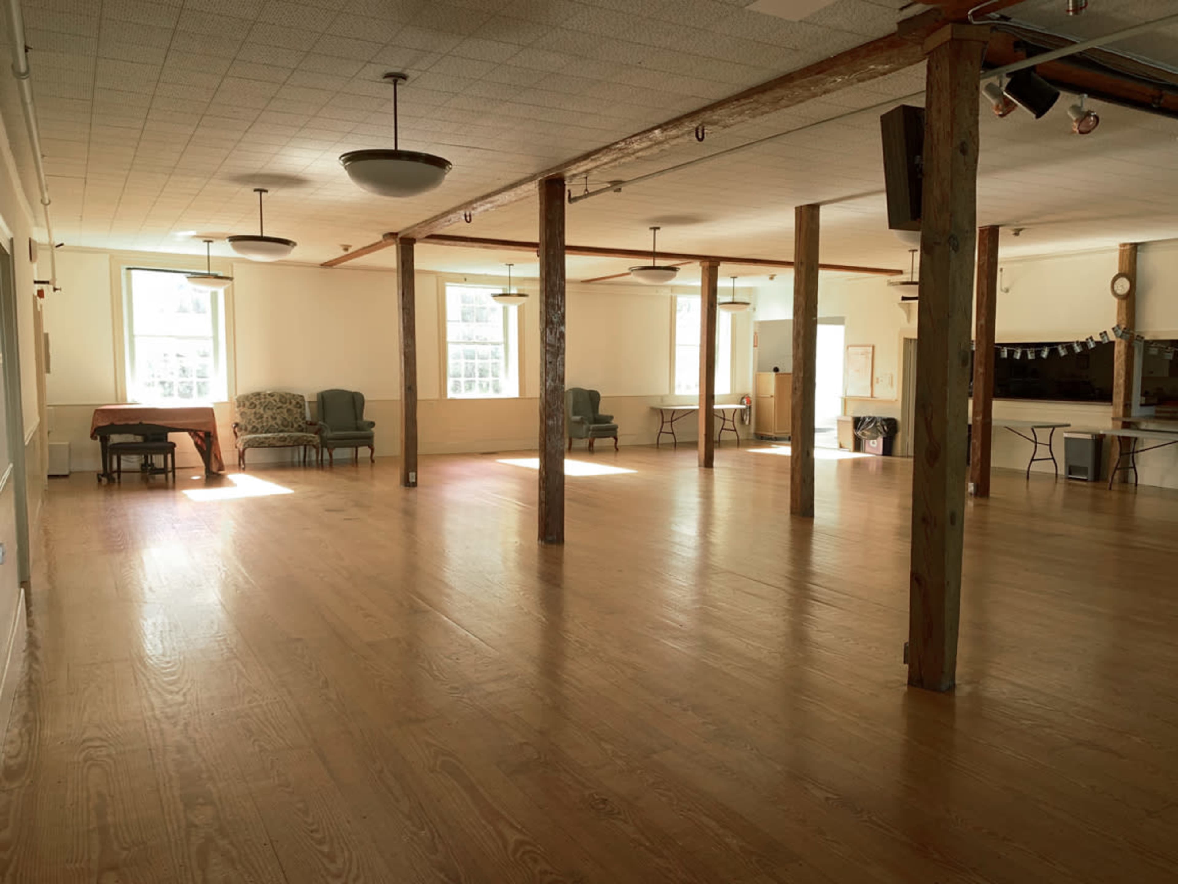 A spacious, well-lit community hall with wooden flooring and several tables and chairs arranged throughout.