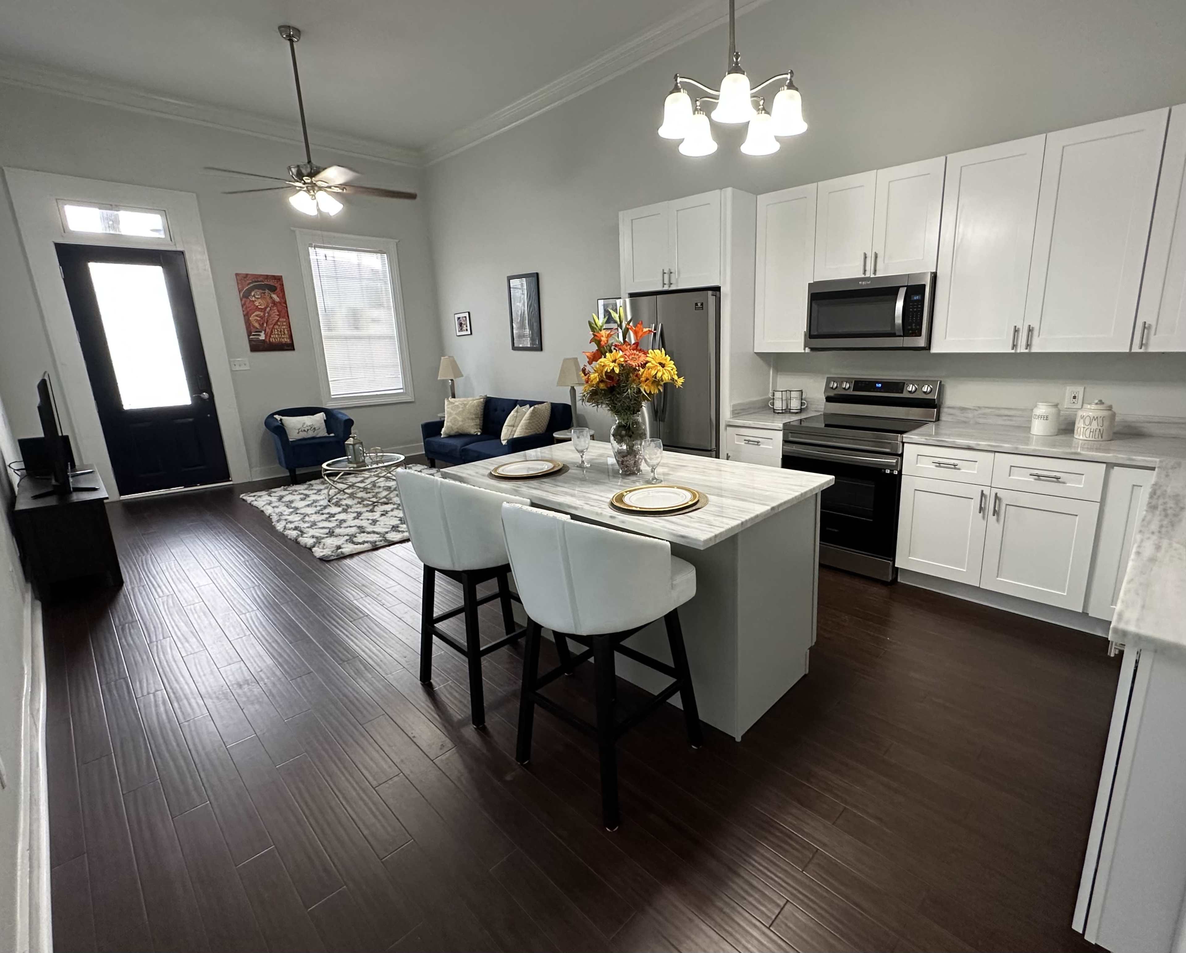 A modern kitchen and living area feature a central island with bar seating, stainless steel appliances, and a seating arrangement with a sofa and a decorative rug.