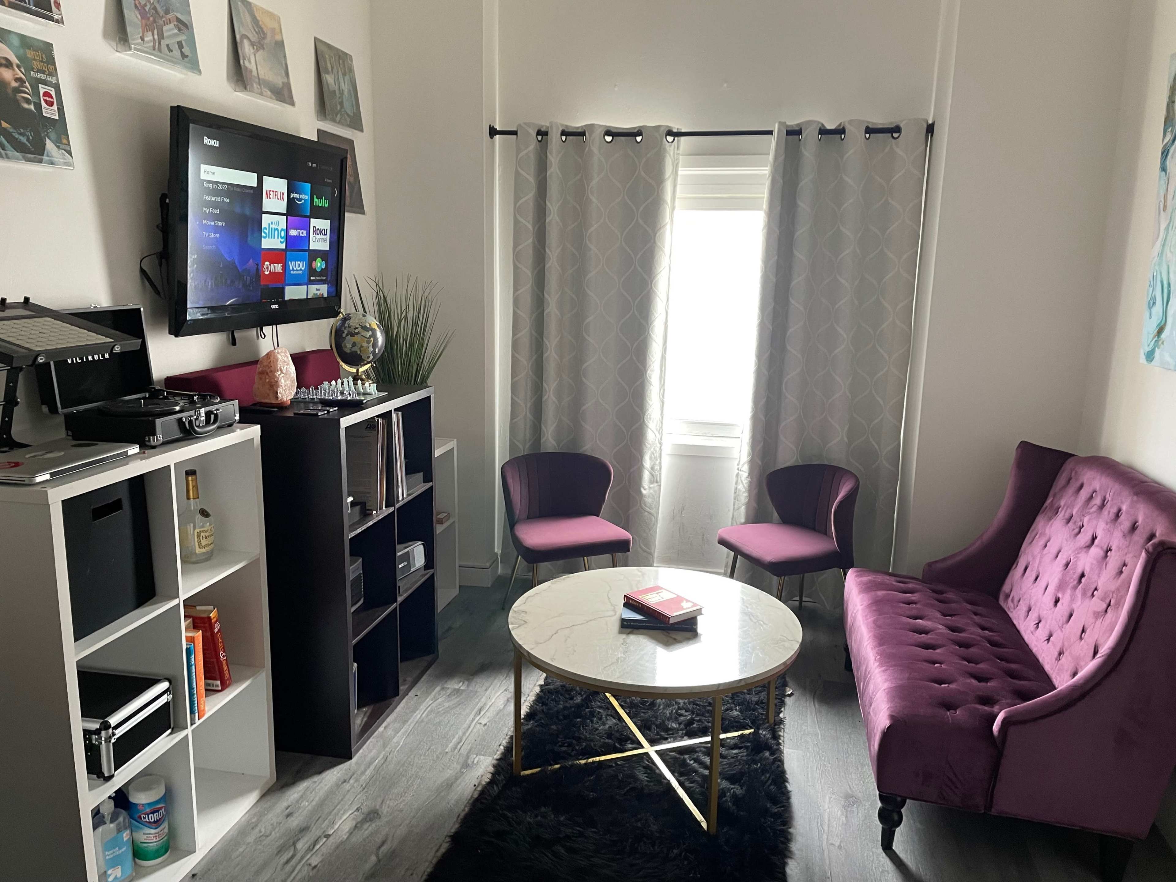 A cozy living room features a purple sofa, two chairs, a round coffee table, a television mounted on the wall, and a window with curtains.