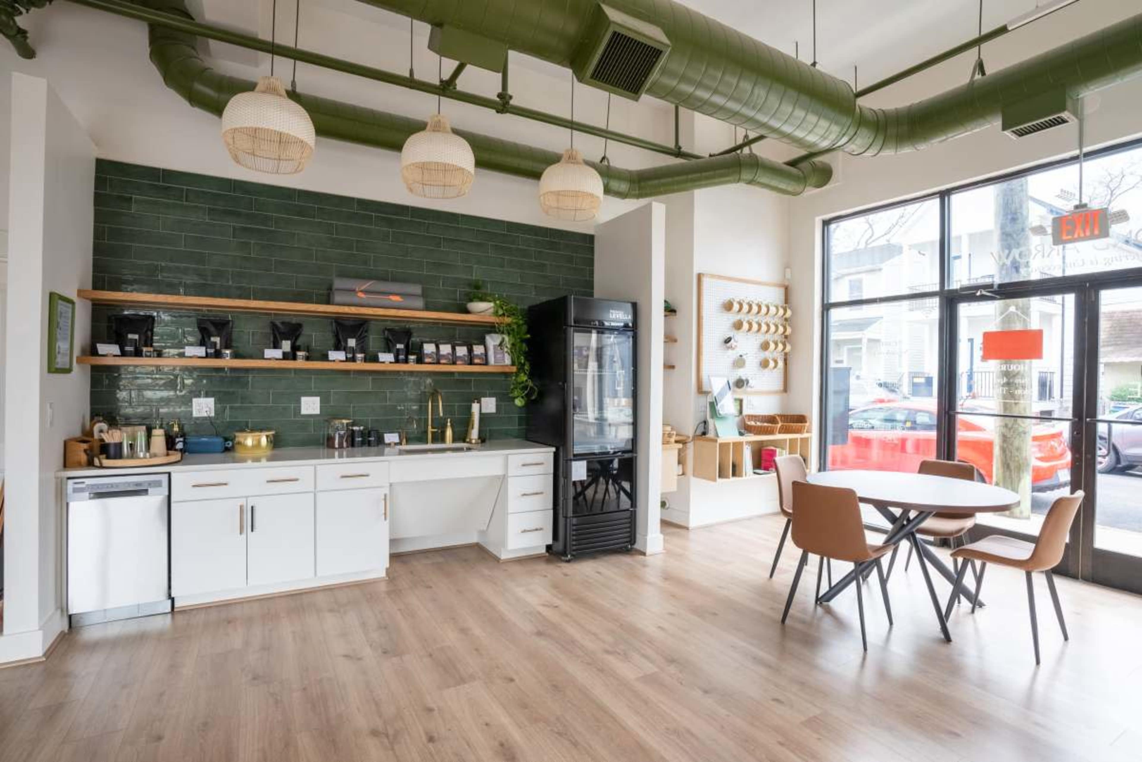 The image shows a modern cafe interior featuring a green tiled wall, wooden shelves stocked with various items, a refrigerator, a coffee station, and a round table surrounded by chairs.