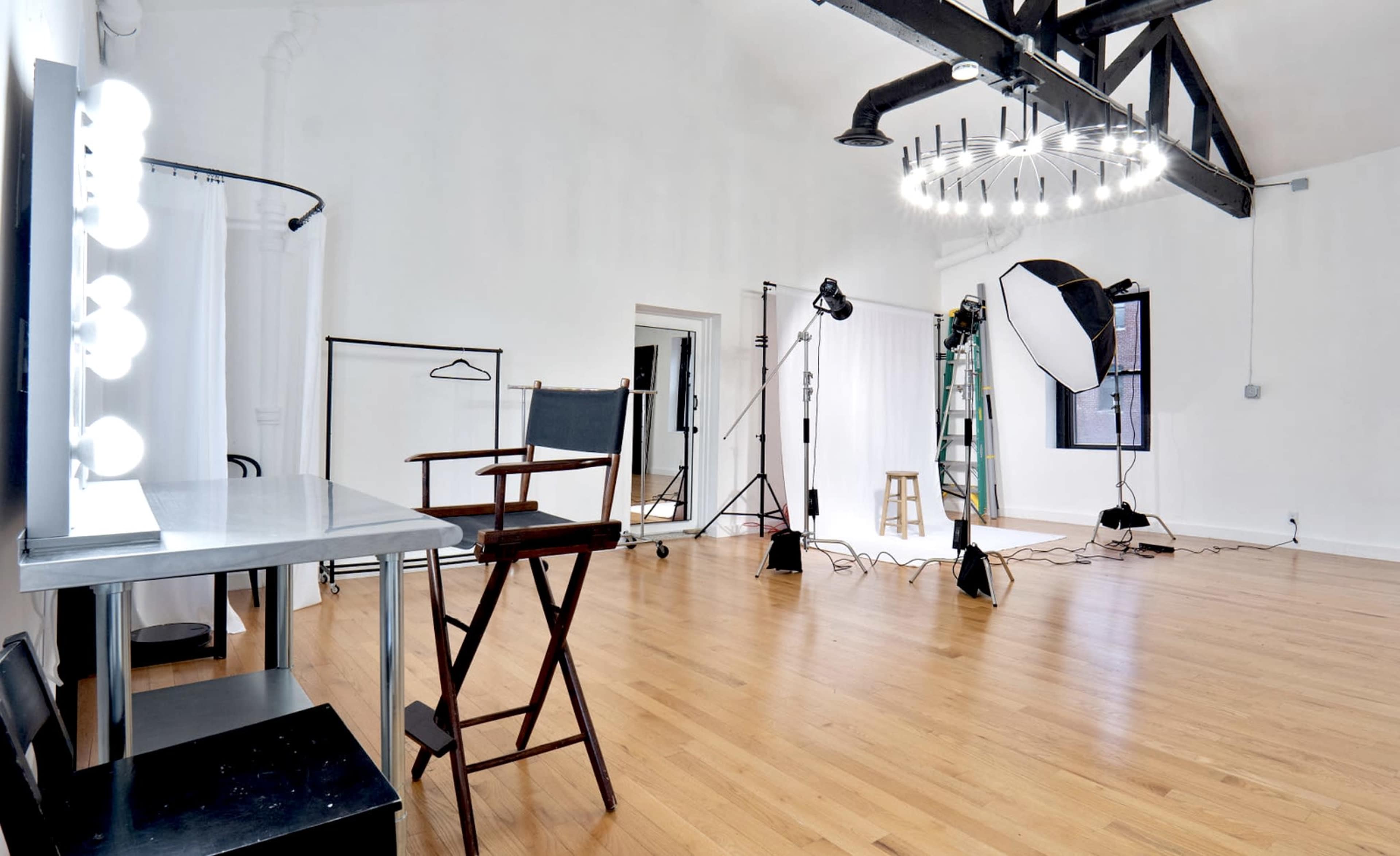 The image shows a brightly lit photography studio with wooden flooring, equipped with softboxes, a makeup mirror, and a director's chair.