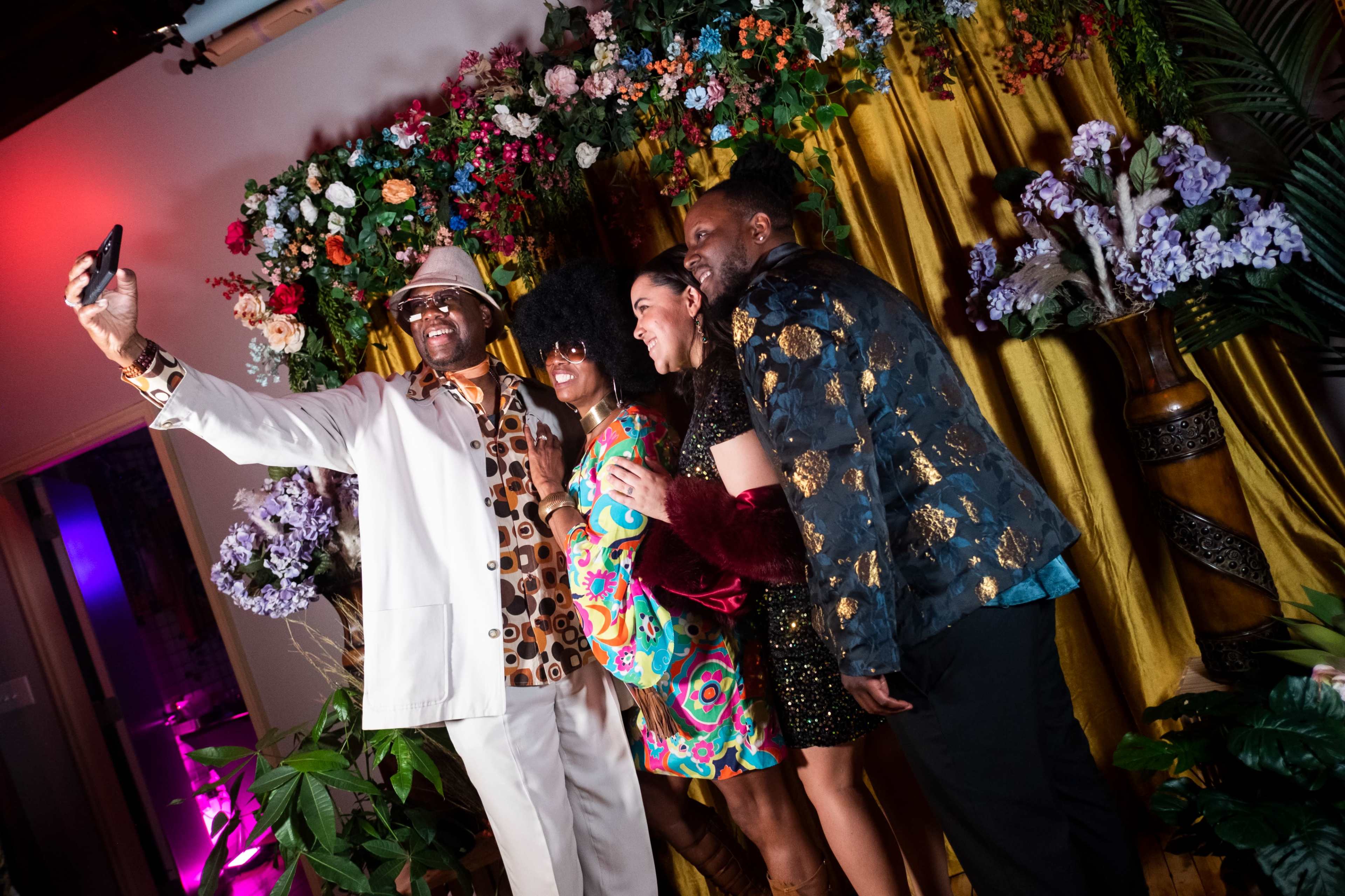 Four people take a selfie in front of a floral backdrop adorned with colorful flowers and greenery.