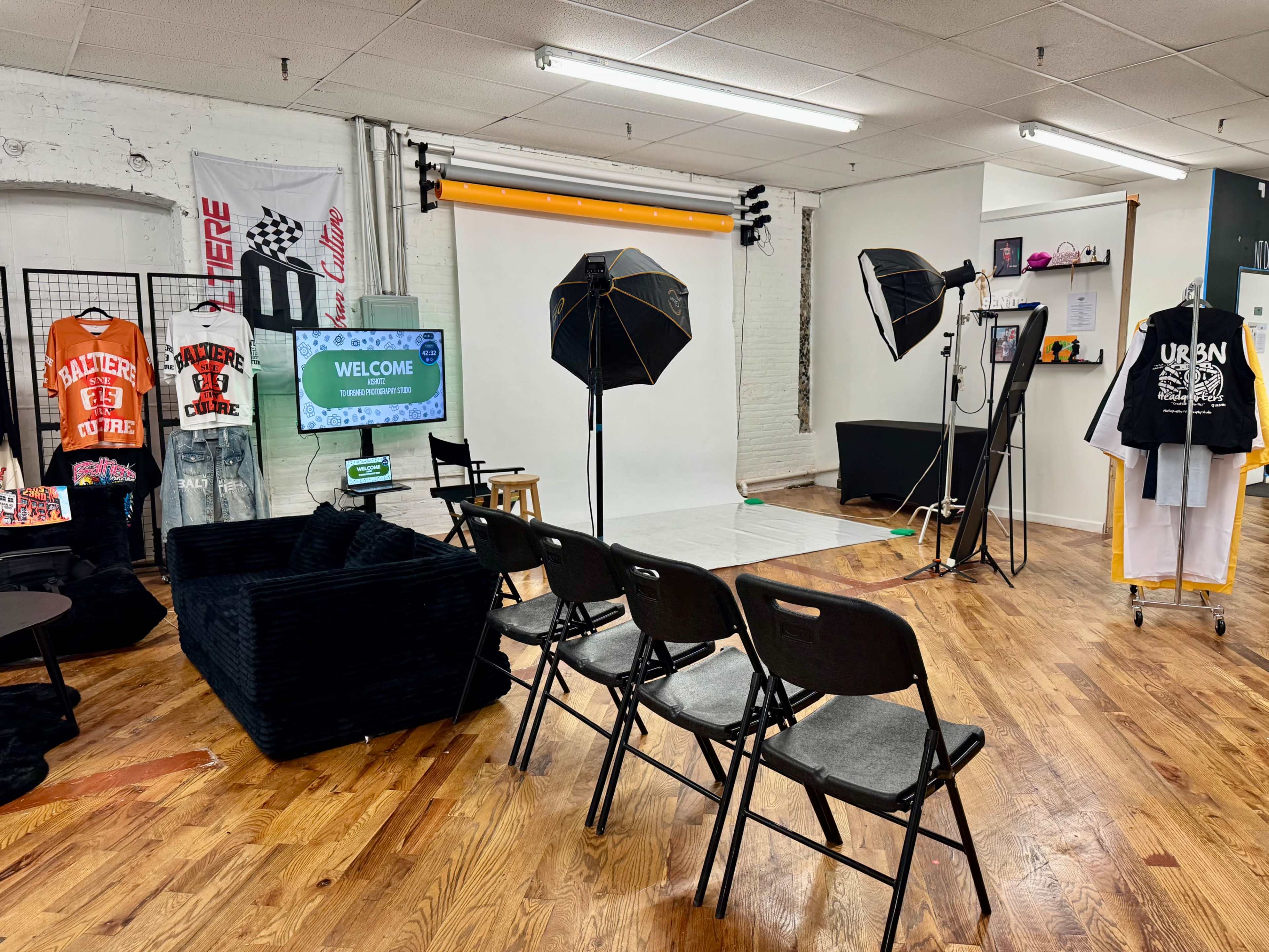 The image shows a spacious studio setup with a white backdrop, lighting equipment, and seating arranged in front of a screen displaying a welcome message.