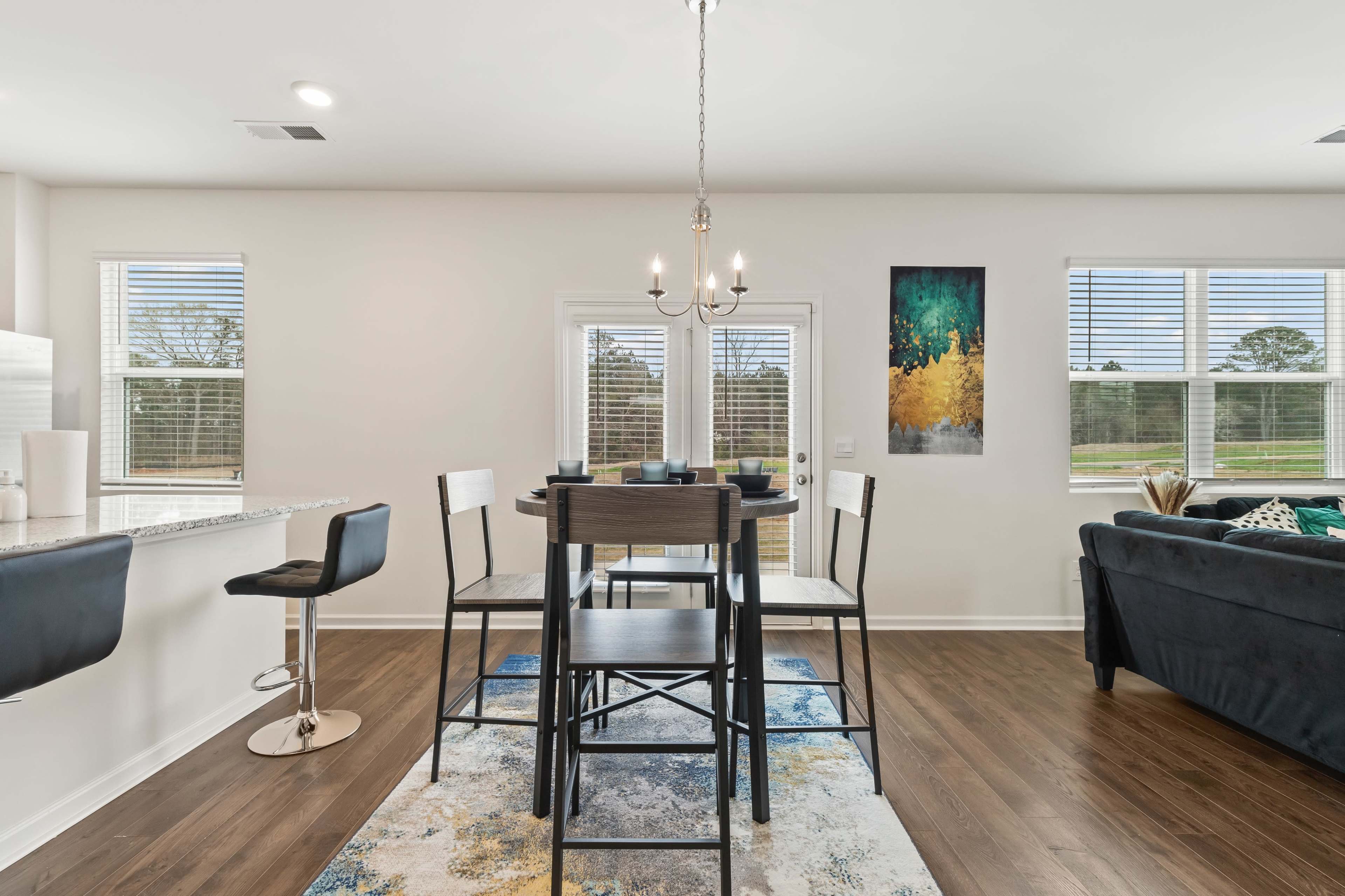 A dining area features a black table and chairs, a chandelier above, and large windows overlooking a backyard.