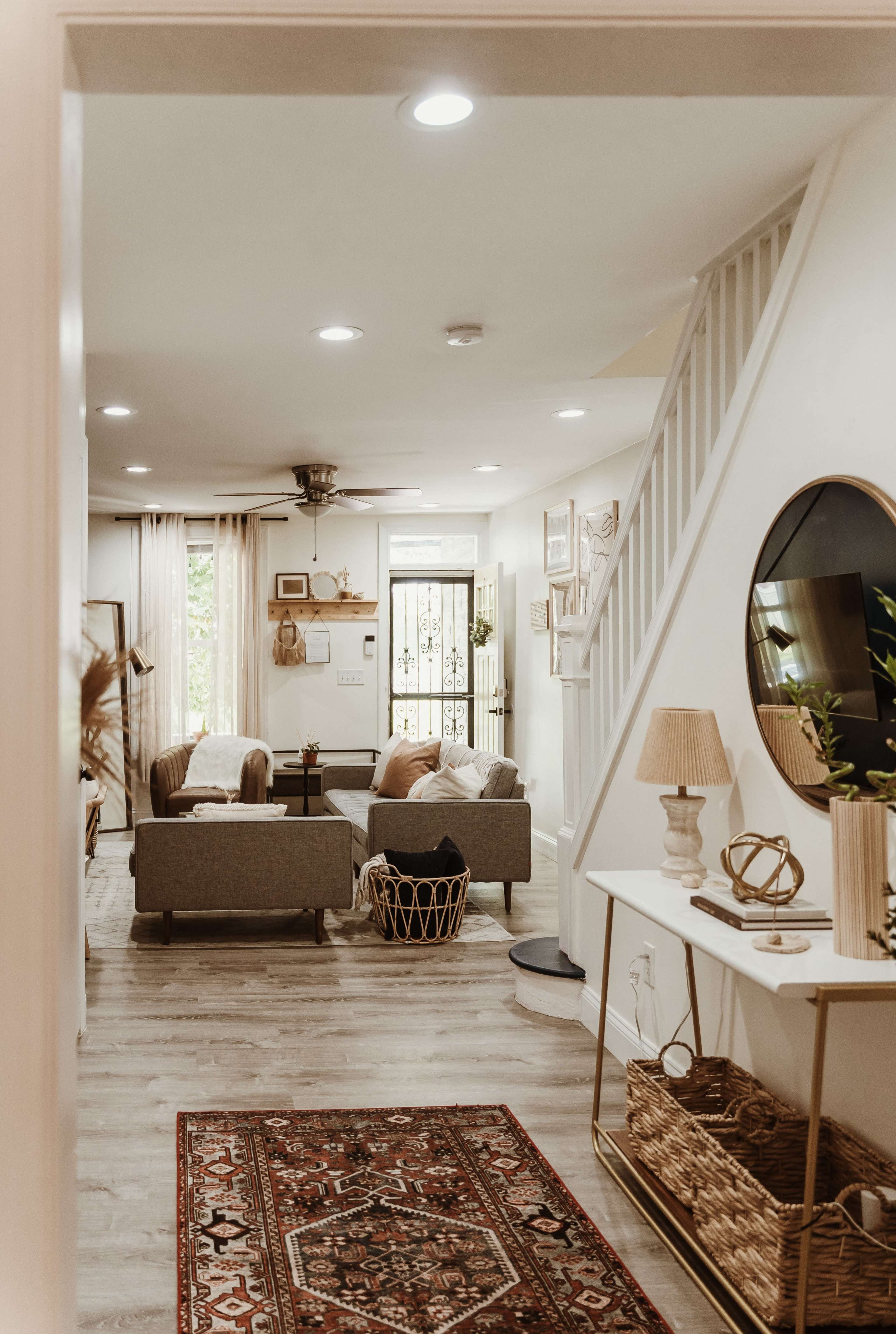 The image shows a well-lit living room featuring gray sofas, a decorative rug, and a staircase, with a view from a hallway.