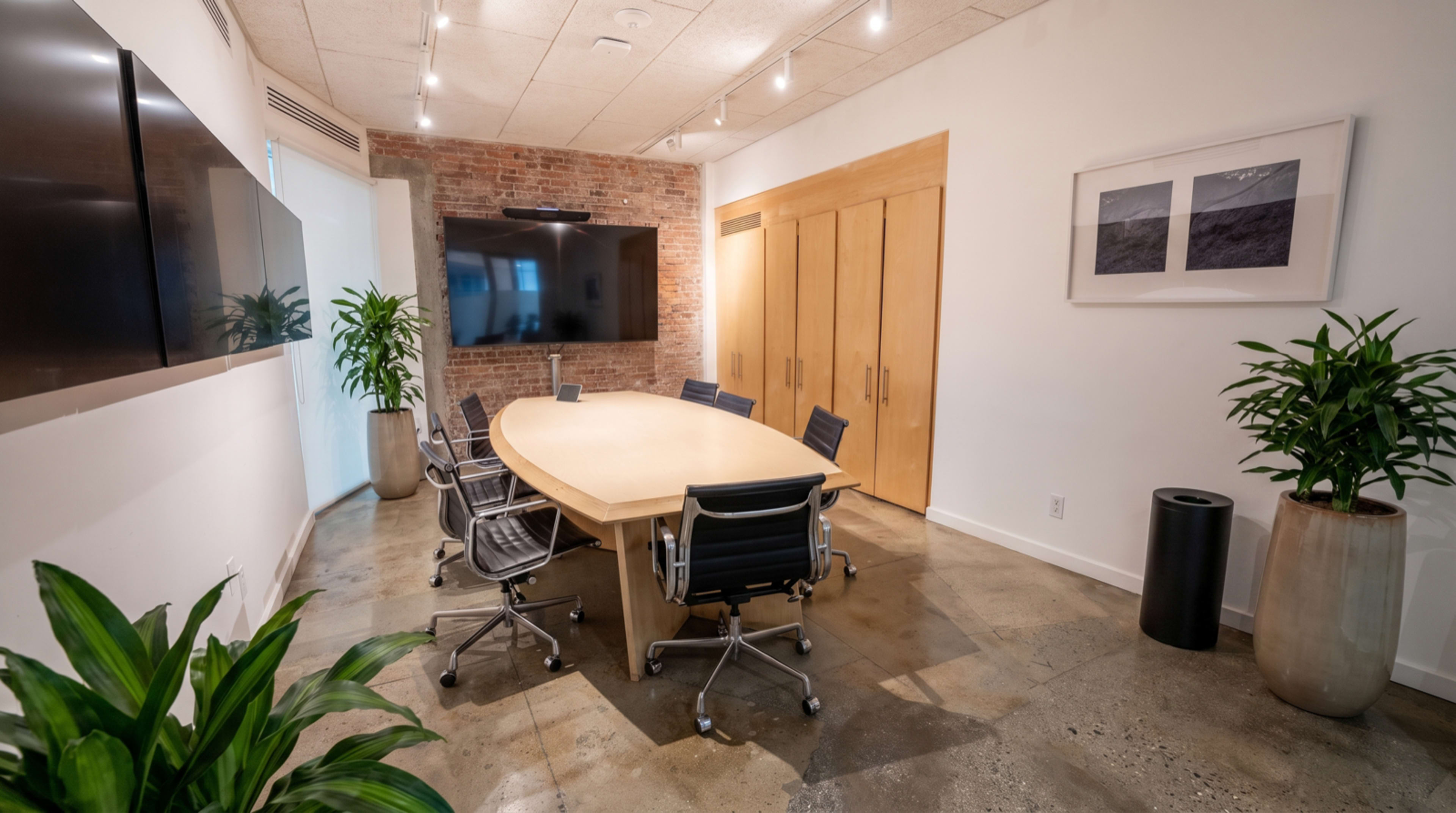 A modern conference room features an oval wooden table surrounded by black leather chairs, with a large screen mounted on a brick wall.