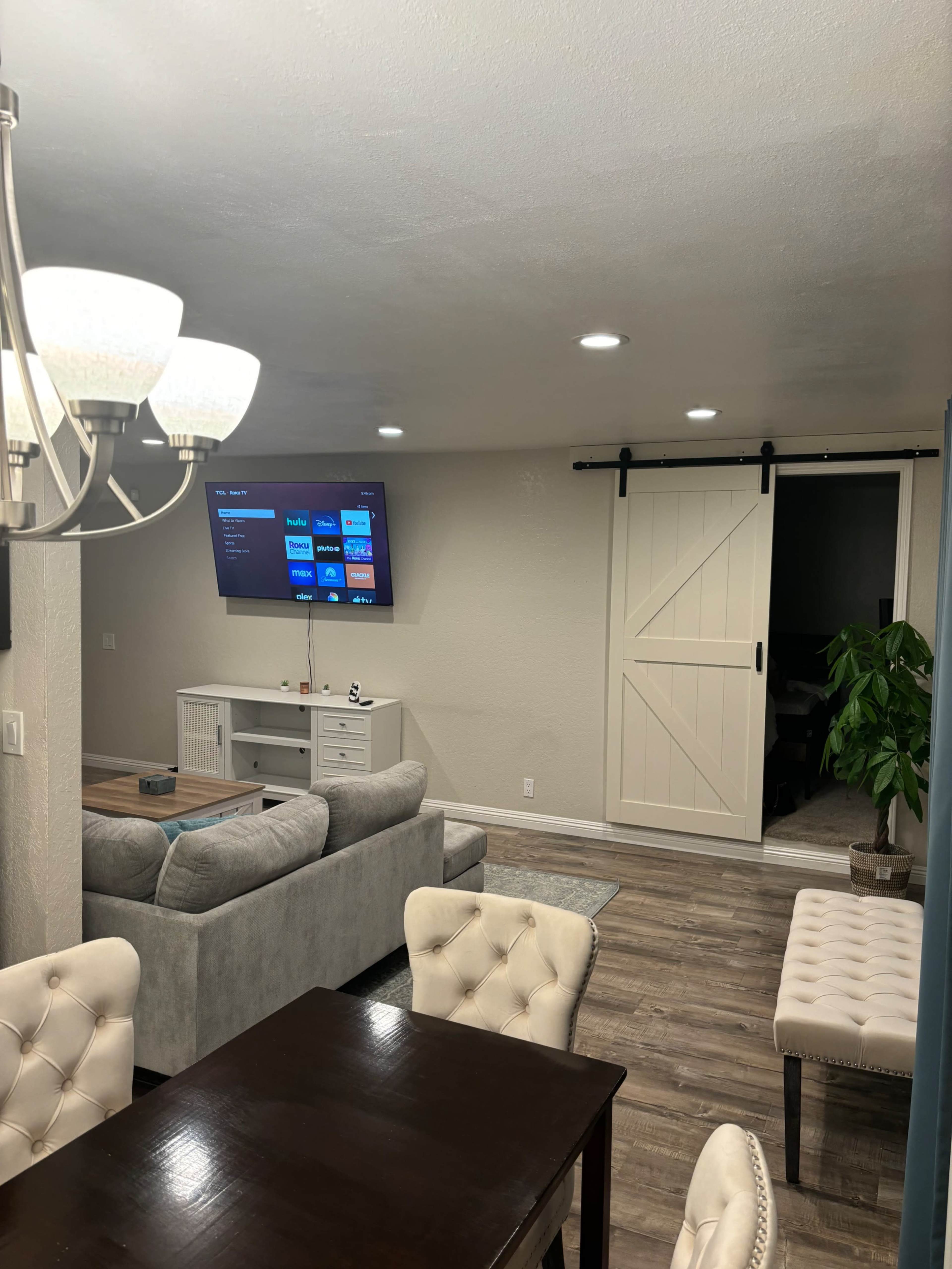 A living room and dining area are visible, featuring a gray couch, a wall-mounted TV, a wooden dining table, and a sliding barn-style door.