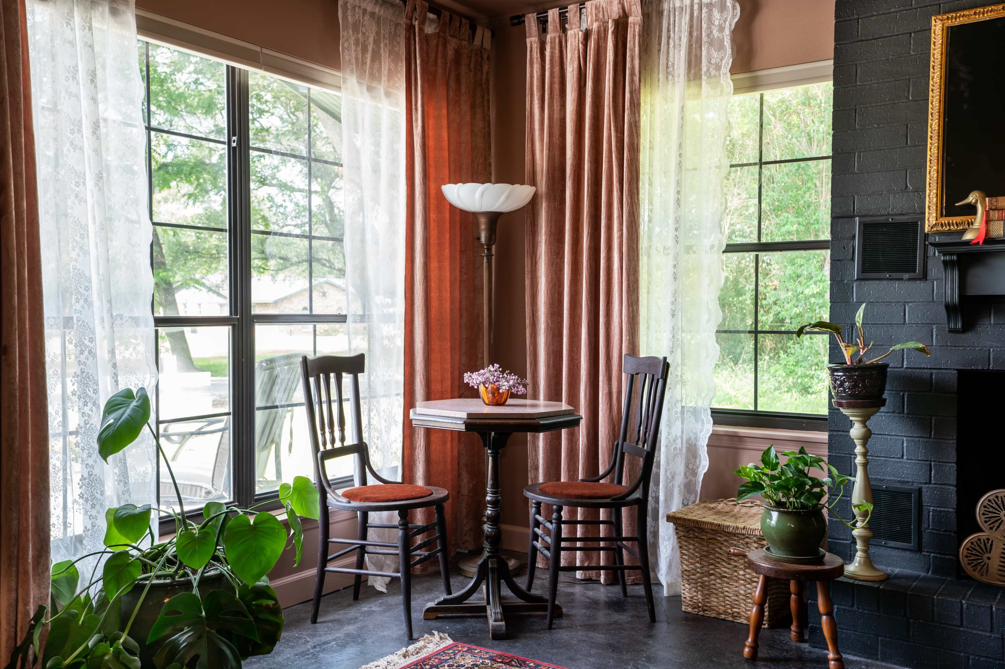 A small table with two chairs sits beside large windows draped with sheer and curtain panels, surrounded by indoor plants and a floral arrangement.