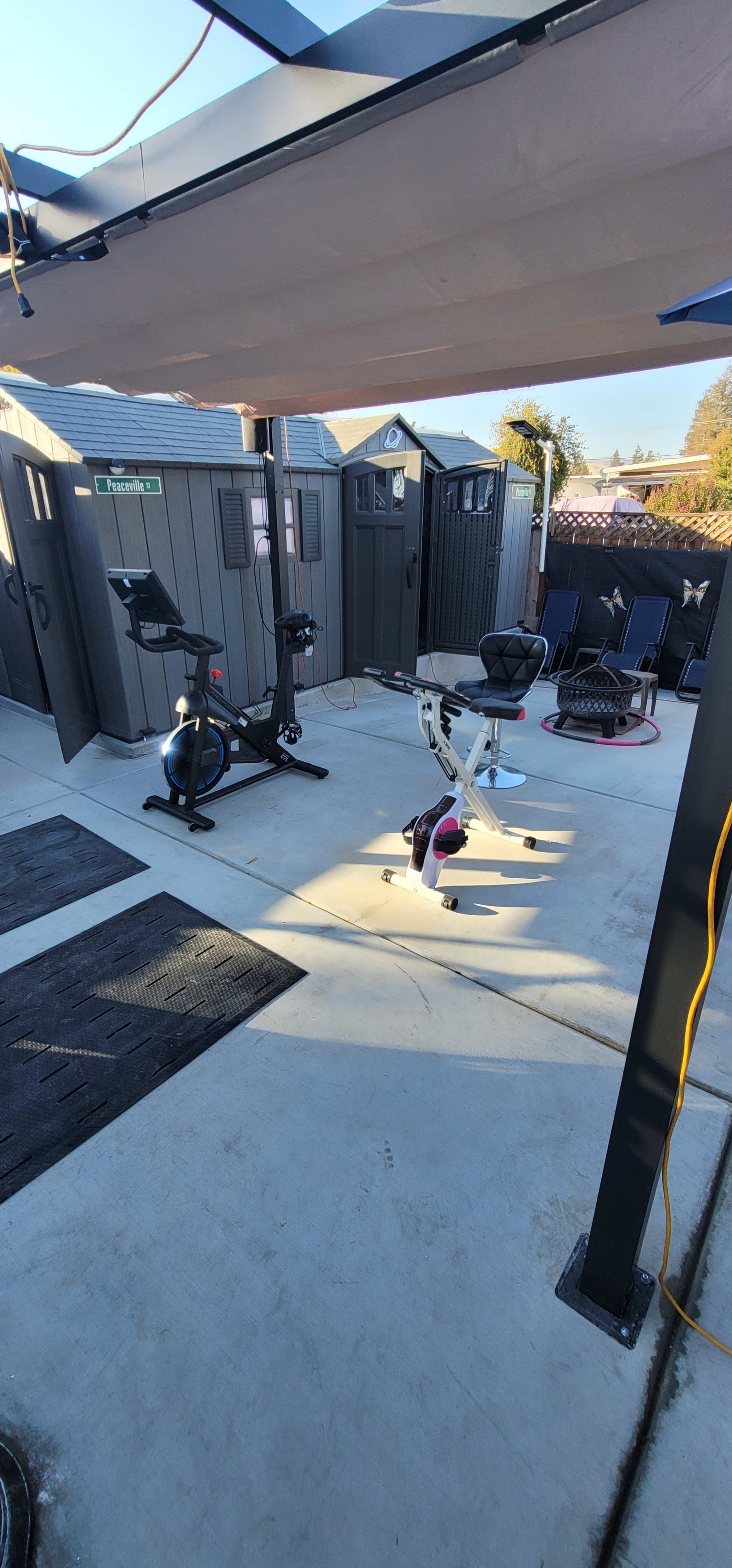 A home gym setup outside, featuring exercise equipment, including a stationary bike and a multi-use exercise bench, on a concrete floor.
