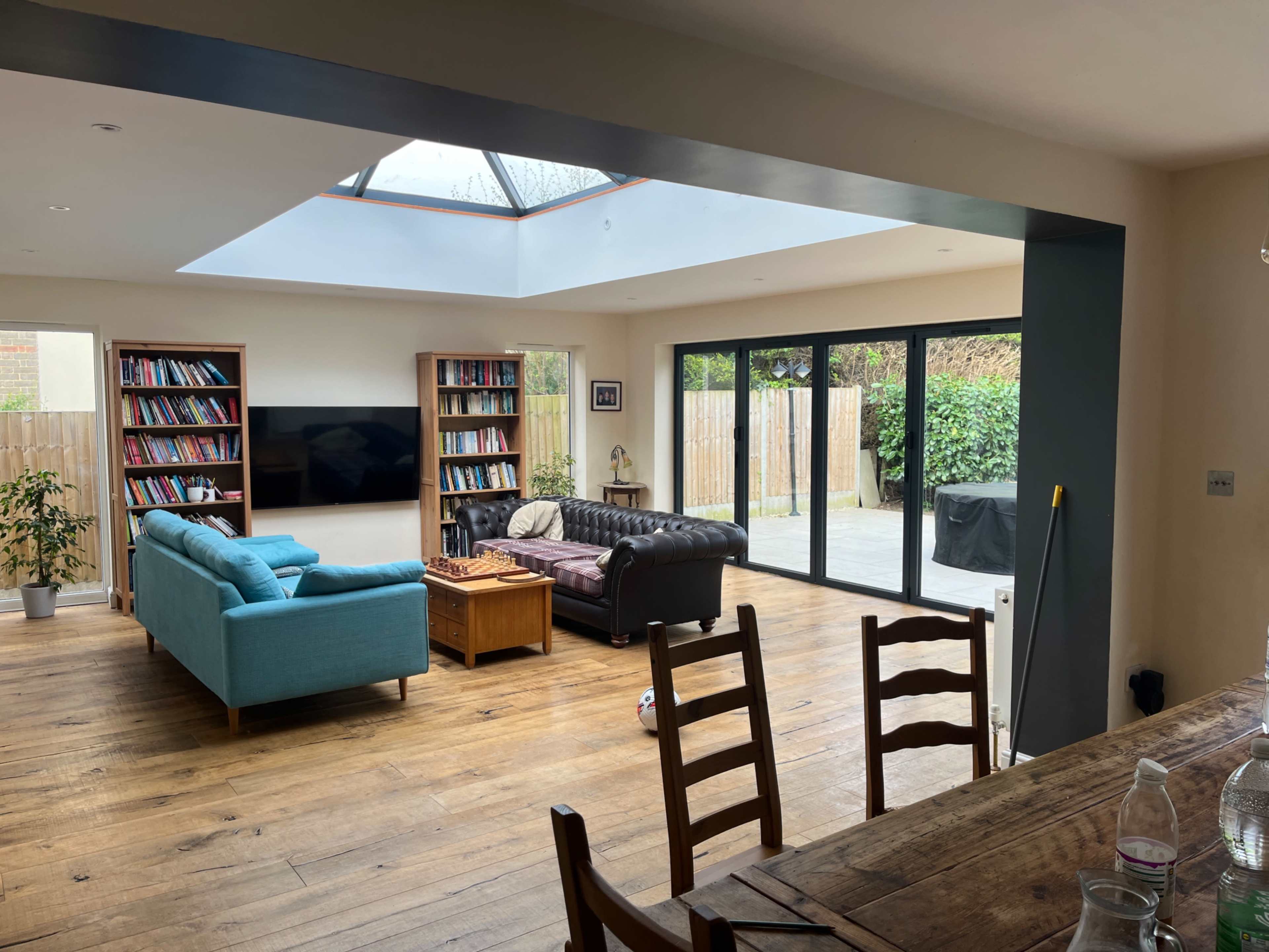 The image shows a spacious living area with a light blue sofa, a dark brown leather sofa, a wooden coffee table, and bookshelves, all illuminated by a skylight.
