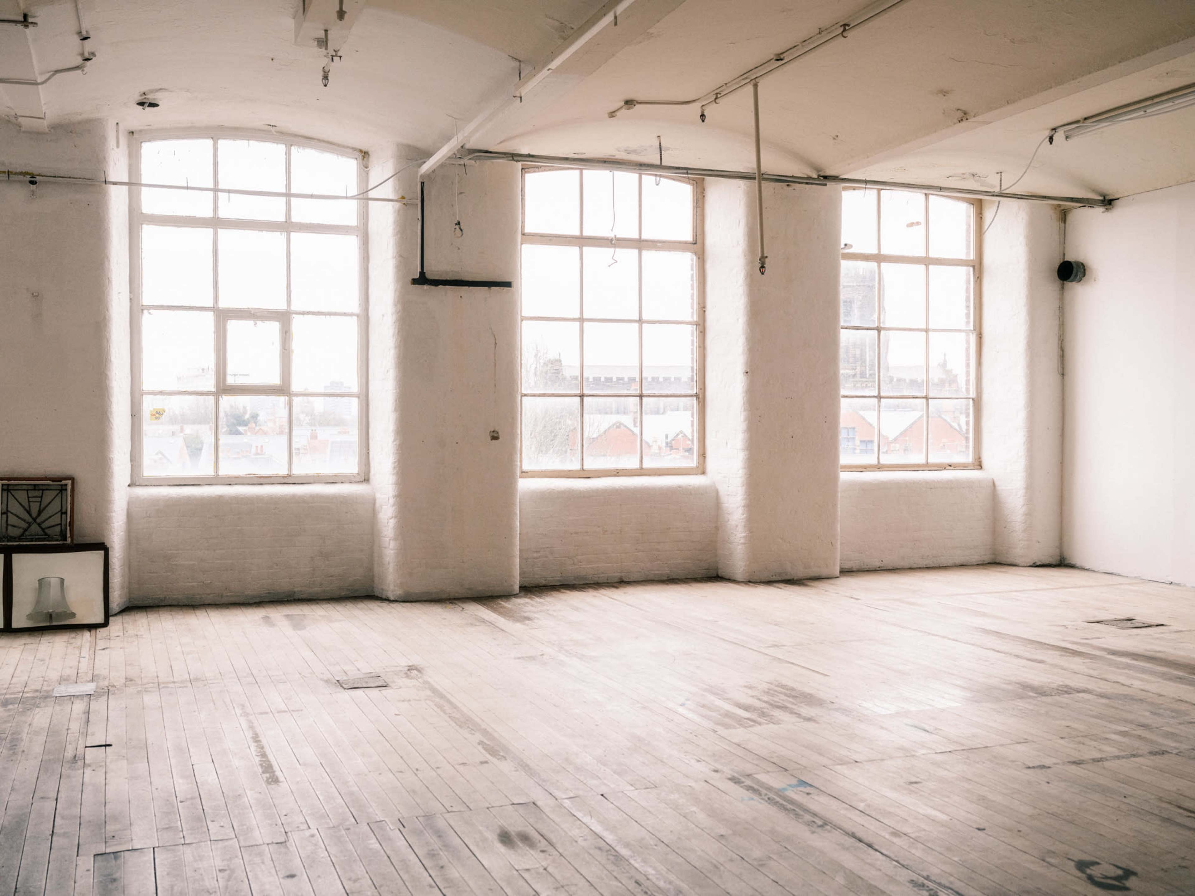 The image shows a spacious, empty room with large windows that allow natural light to fill the space, and wooden floors contrasting with white walls.