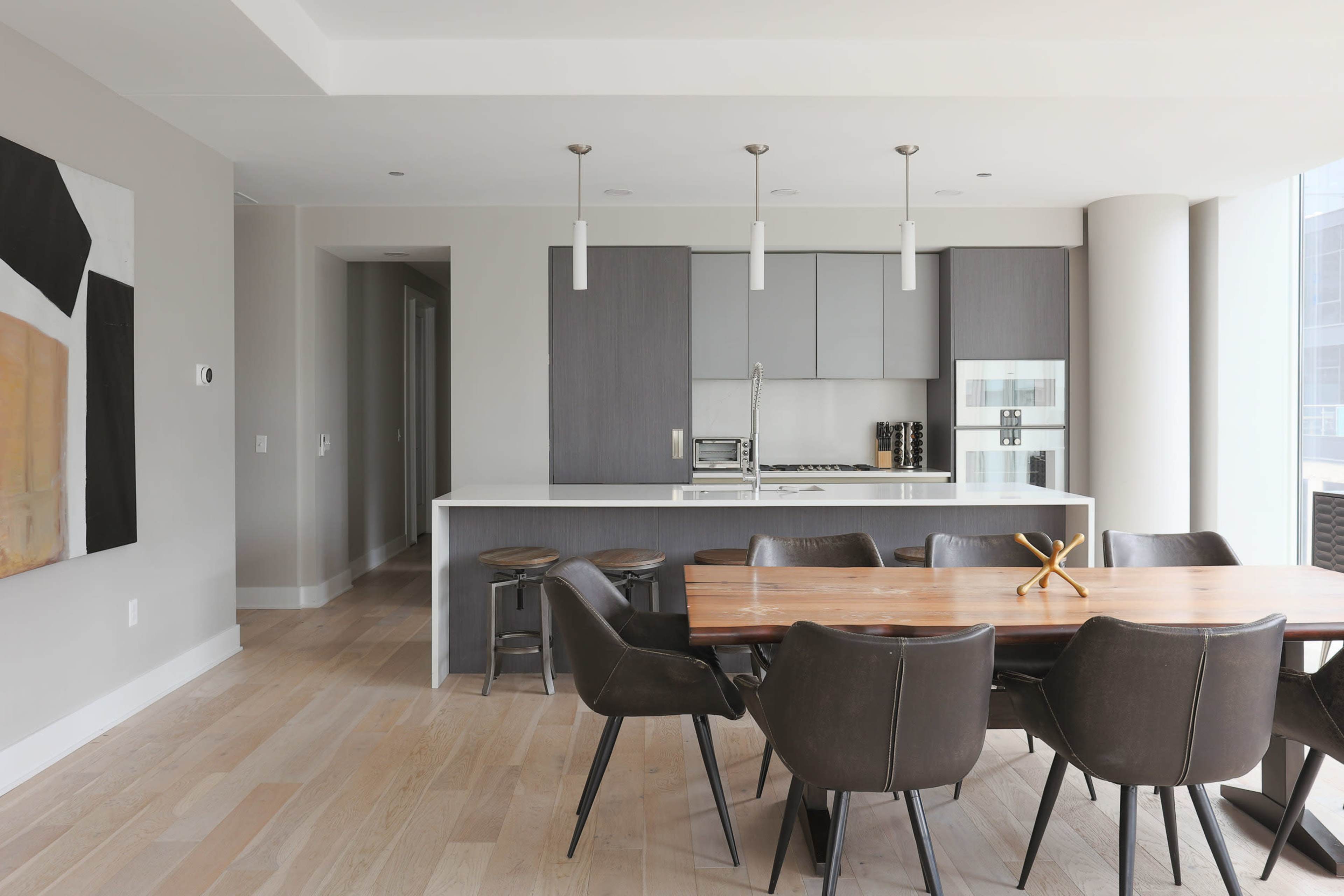 A modern kitchen and dining area features a wooden table surrounded by leather chairs, with sleek cabinetry and a countertop in the background.