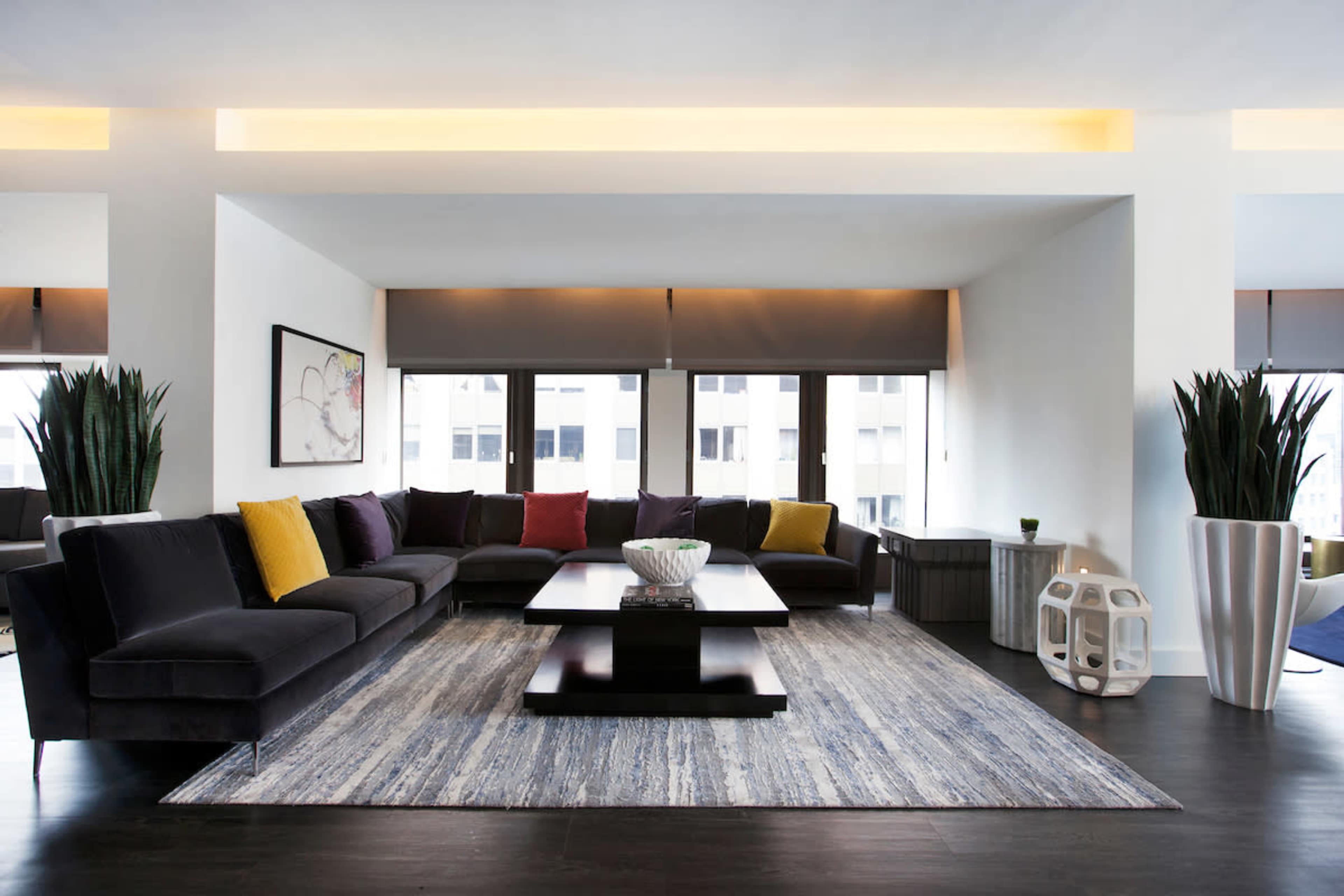 A modern living room features a large dark sofa with colorful cushions, a central coffee table, and a patterned area rug beneath it.