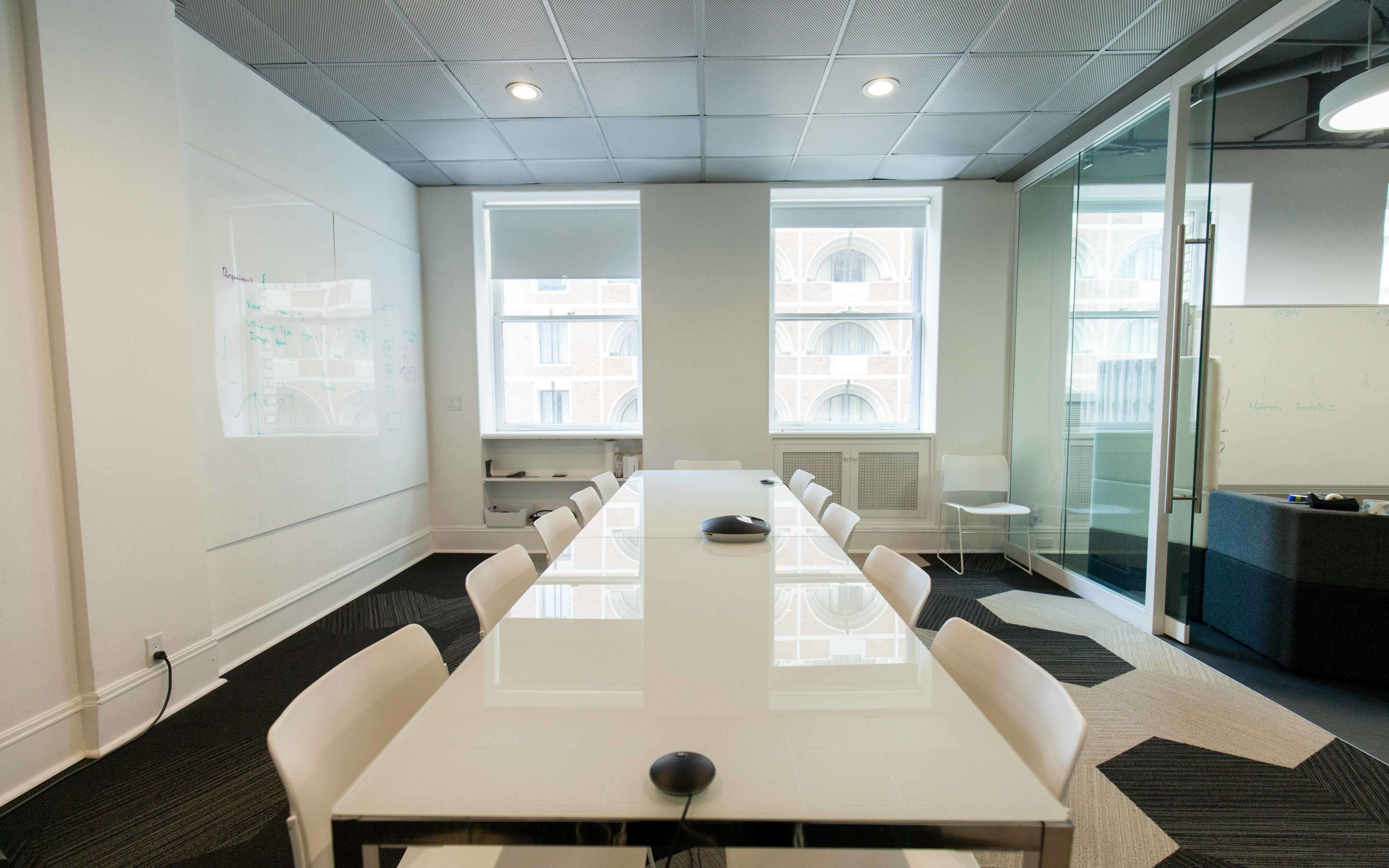 A modern conference room features a long white table surrounded by white chairs, with large windows allowing natural light and a glass wall separating it from another space.