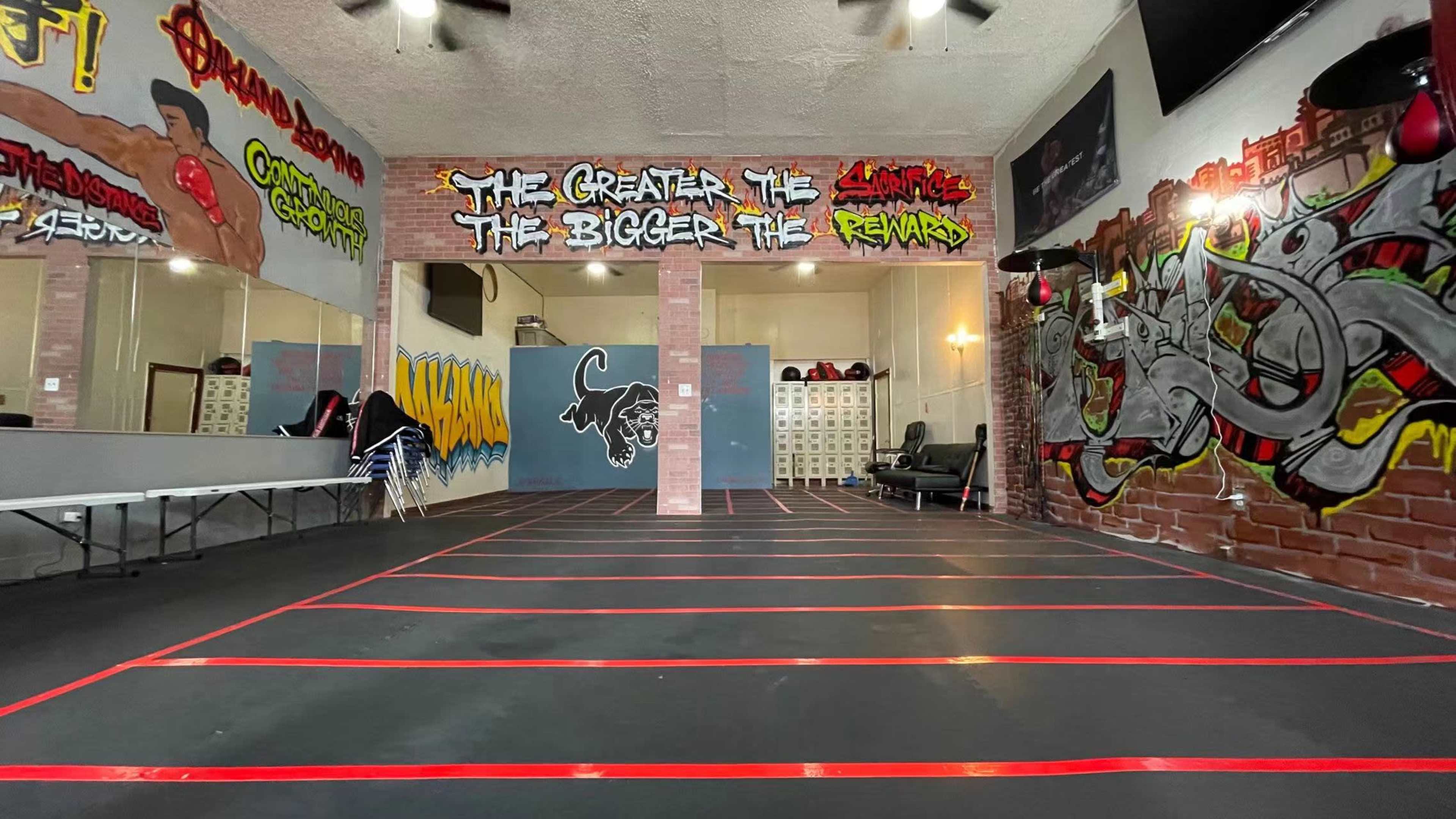 A gym interior with graffiti-covered walls and a spacious open area marked by red tape lines on the black floor.