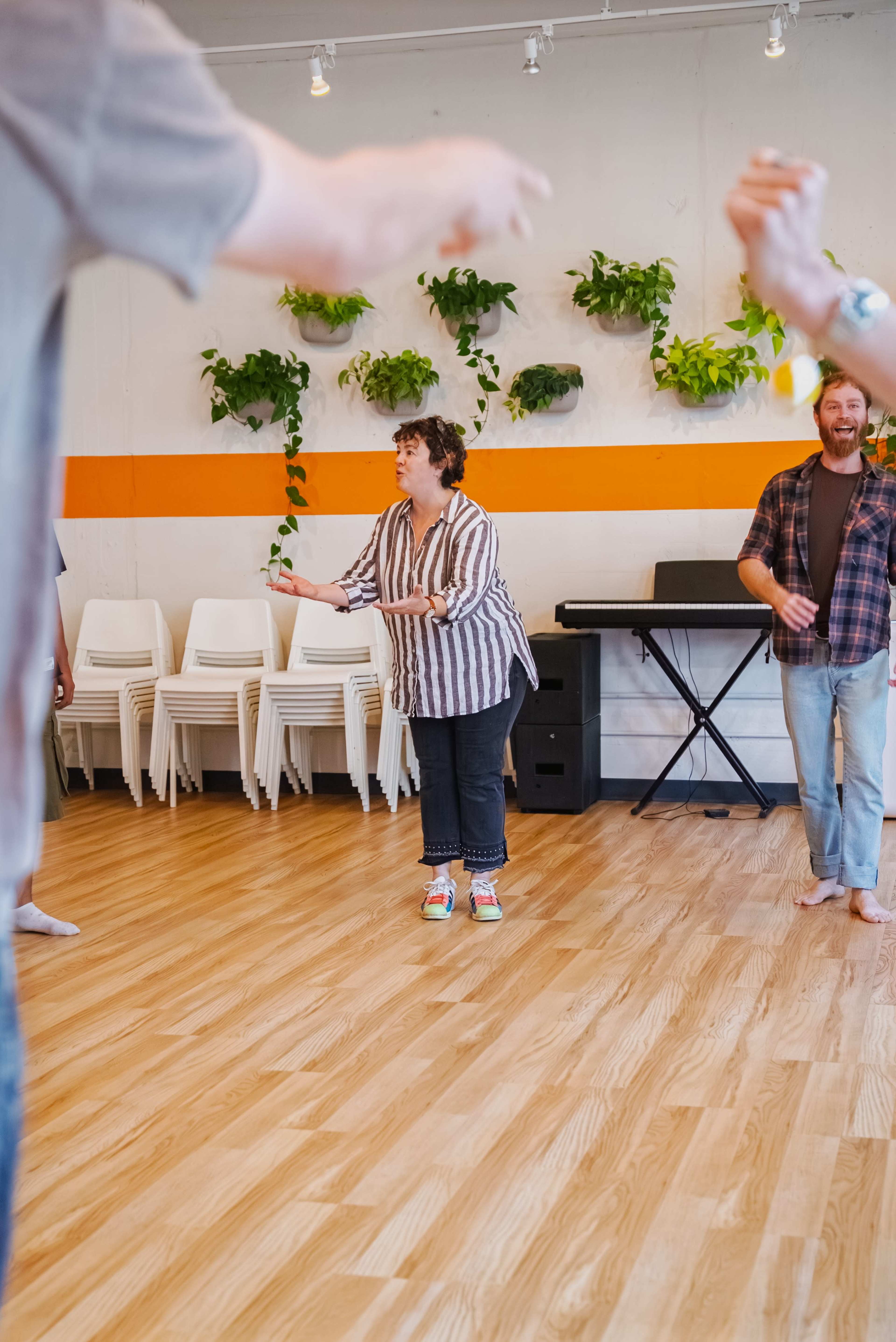 A group of people stands in a spacious room with wooden floors and plants on the wall, while one person in a striped shirt gestures as they interact.