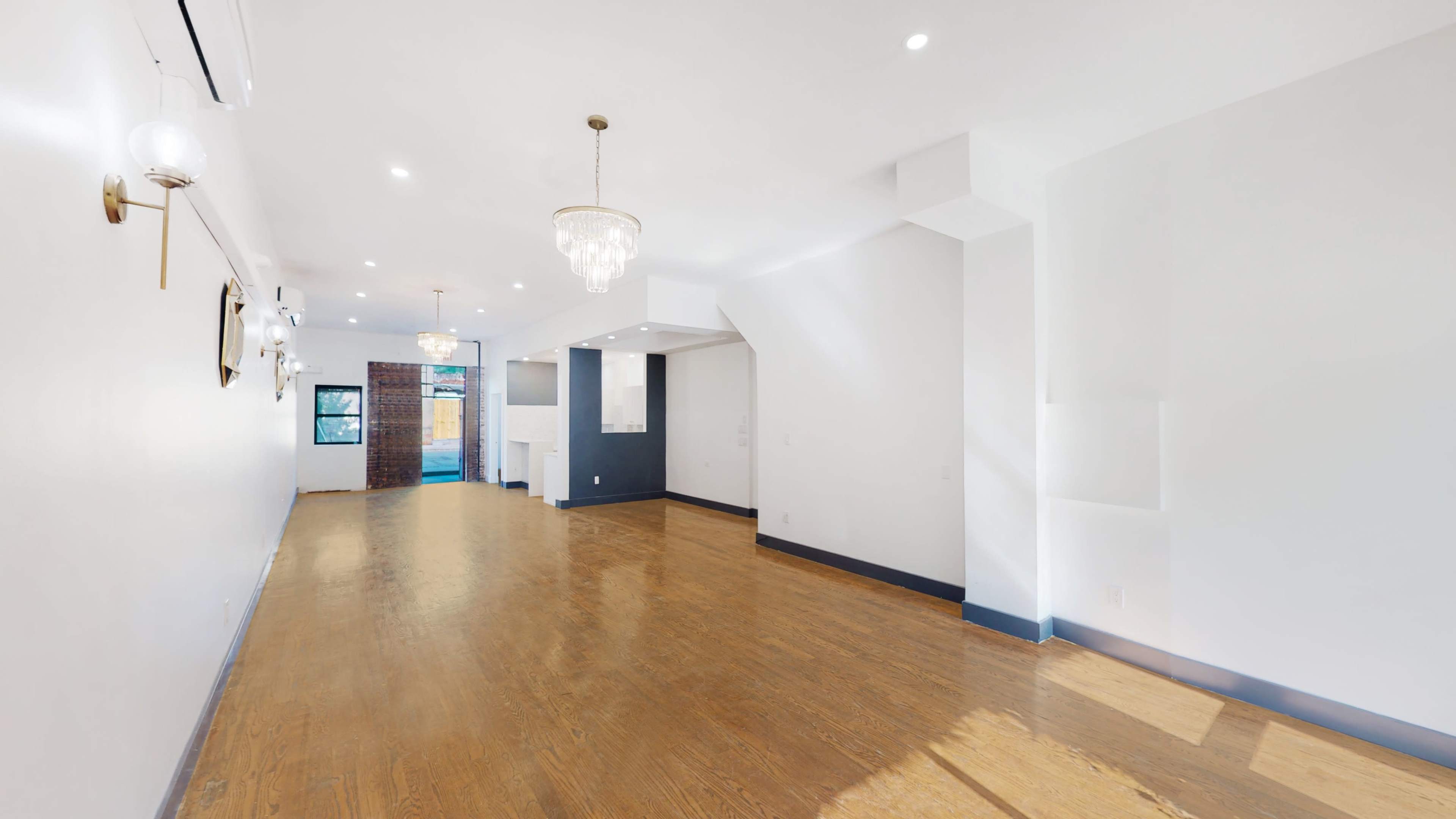 A spacious, well-lit interior room with hardwood flooring, featuring two ceiling chandeliers and a minimalist design.