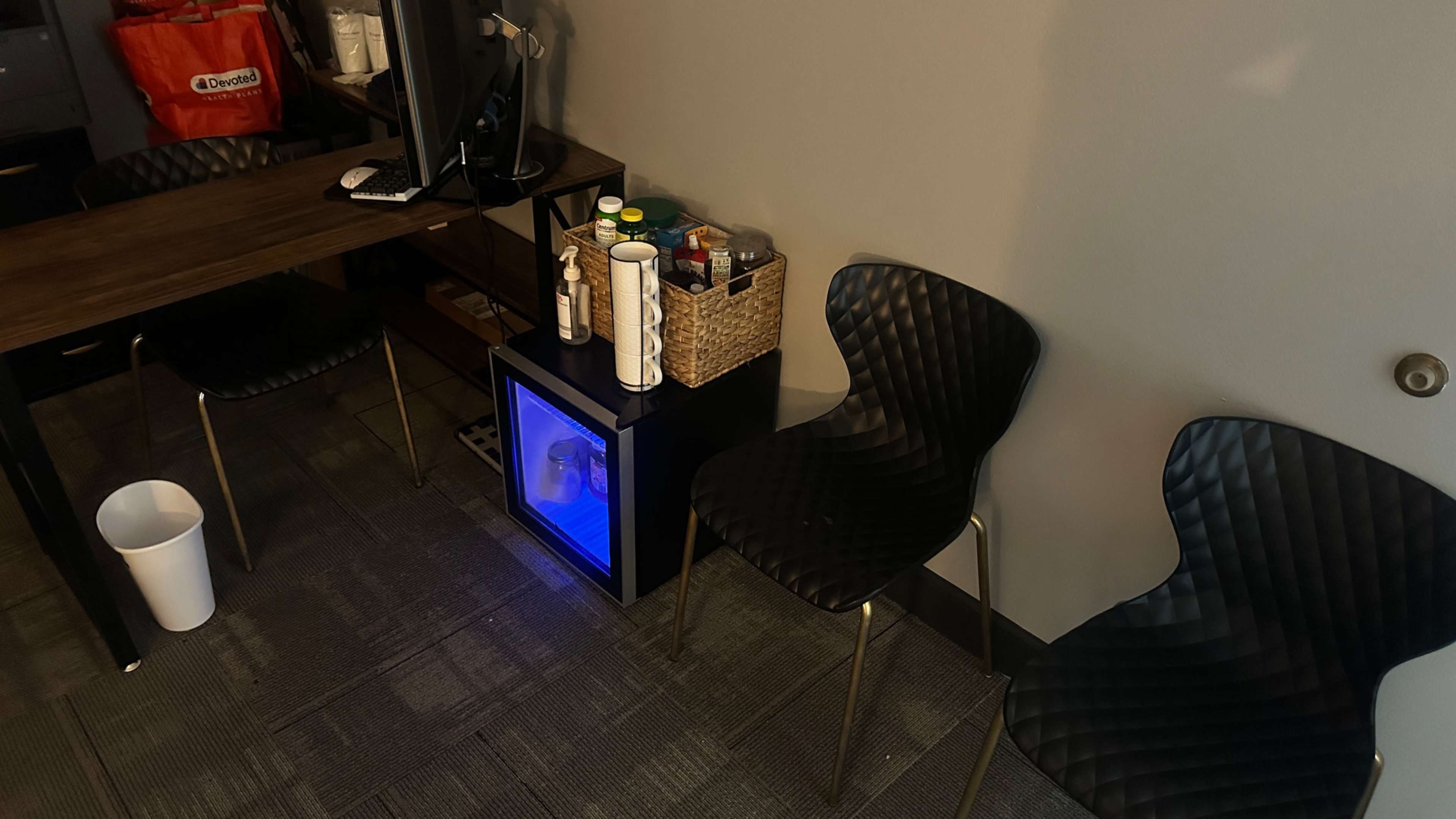The image shows a corner of an office space featuring two black chairs, a small refrigerator with a blue light, and a basket containing various items on a wooden table.