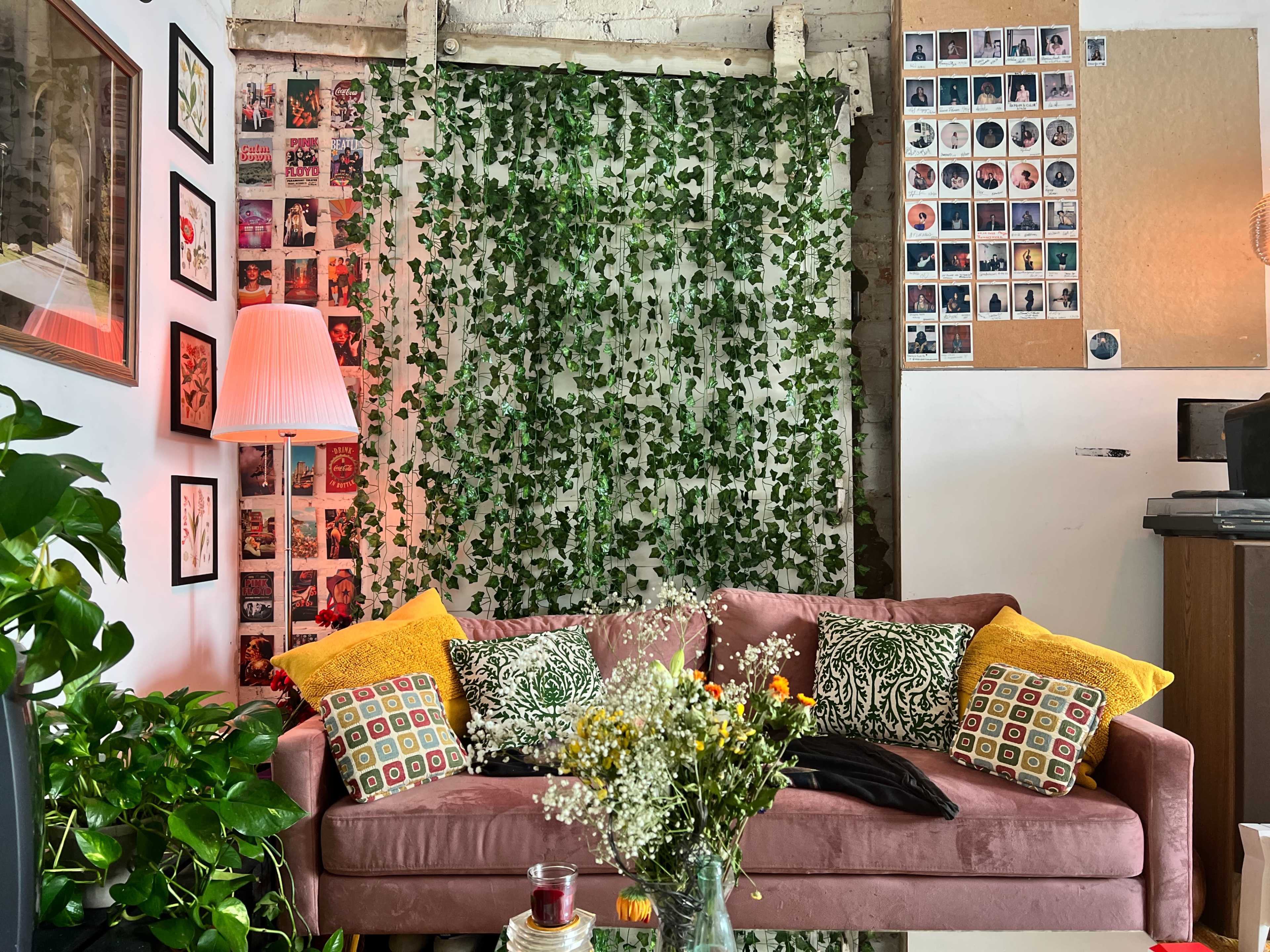 The scene features a pink sofa adorned with patterned pillows, surrounded by greenery and framed photographs on the walls, along with a warm lamp and a vase of flowers on a table.