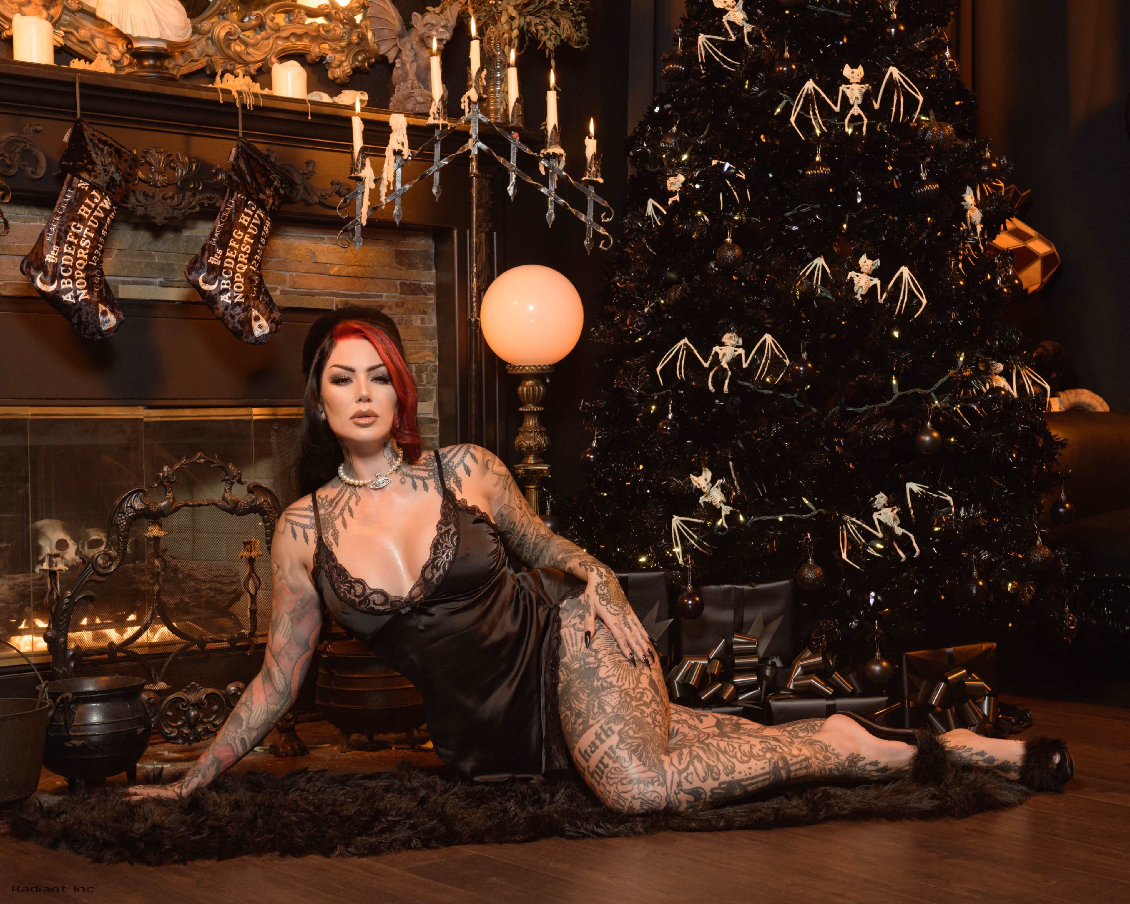 A woman with tattoos poses in front of a decorated black Christmas tree and a fireplace with candles and stockings.
