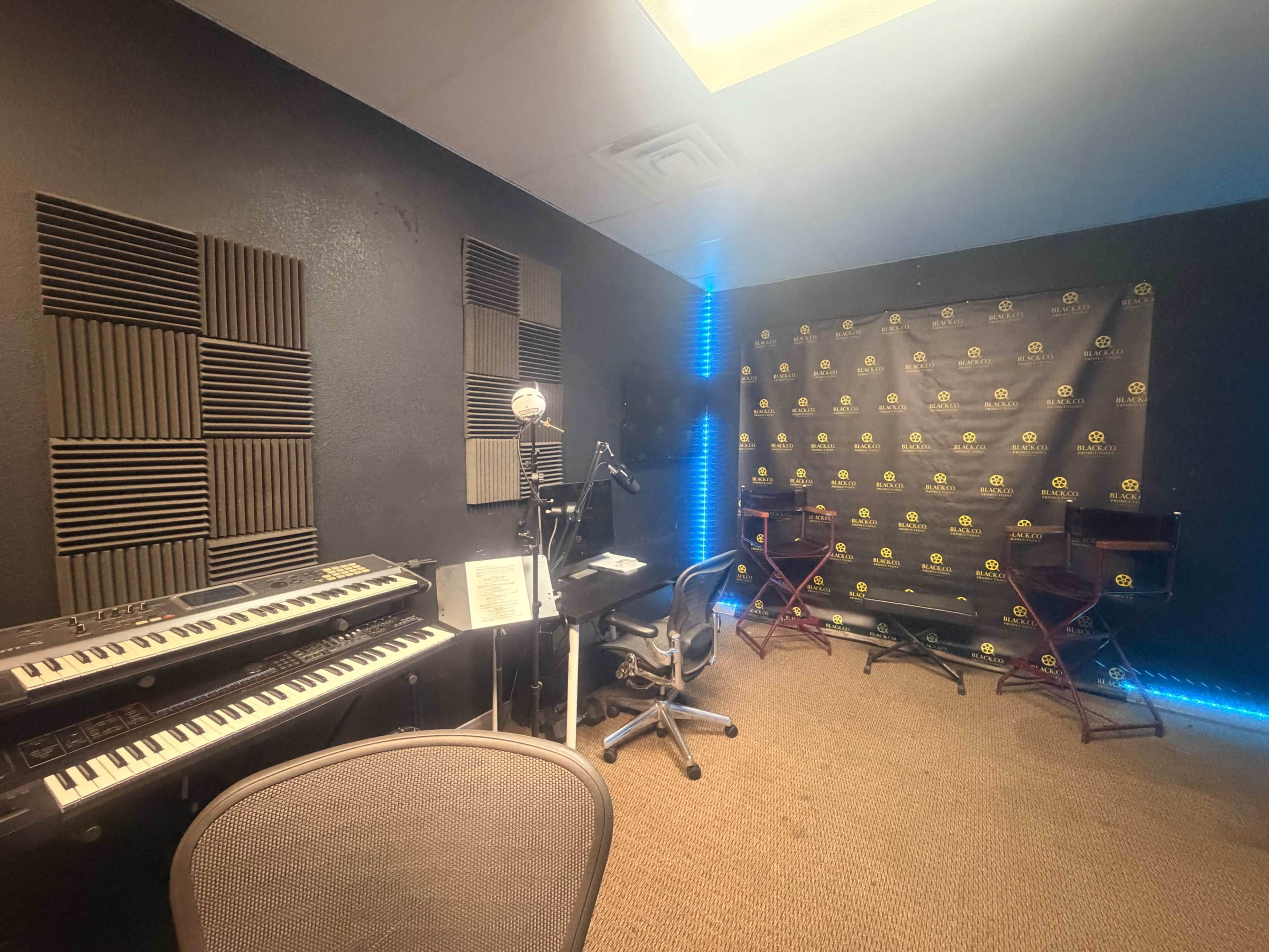 The image shows a music studio featuring a keyboard, microphone, chairs, and a backdrop with logos.