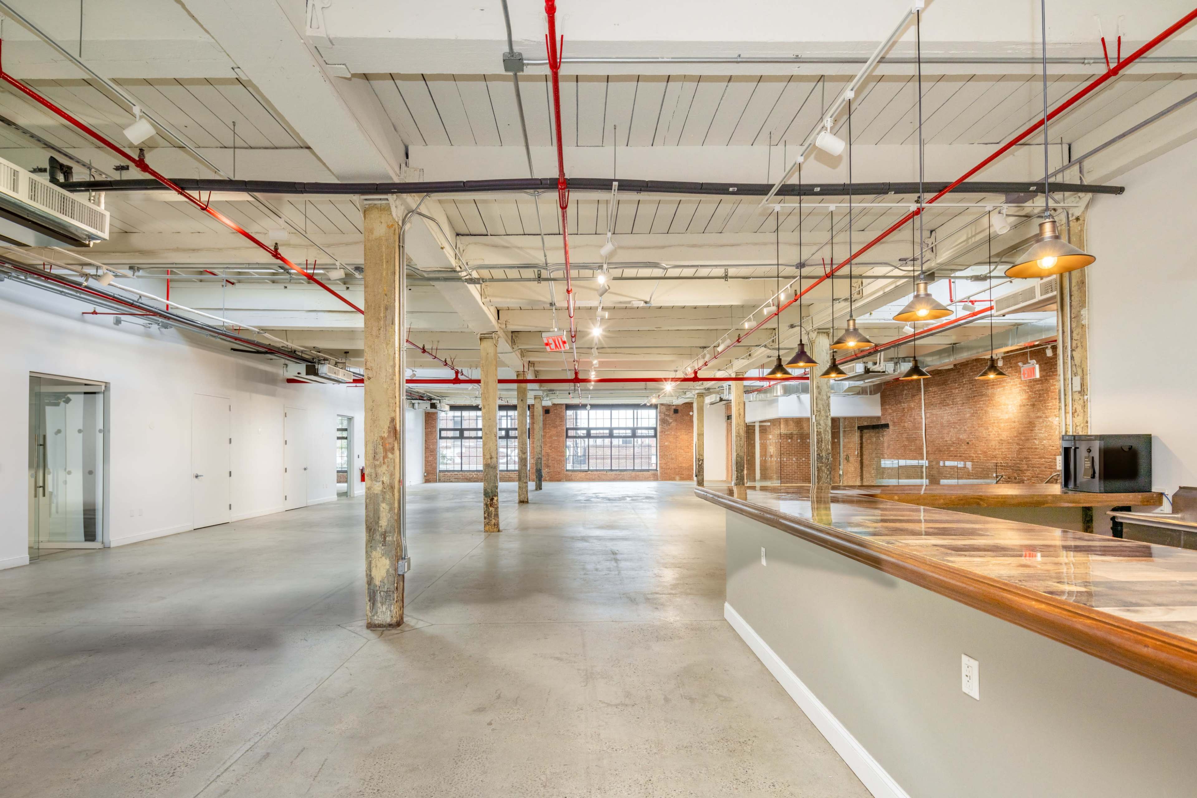 A spacious, industrial-style interior with exposed brick walls, concrete floors, overhead piping, and large windows allowing natural light.