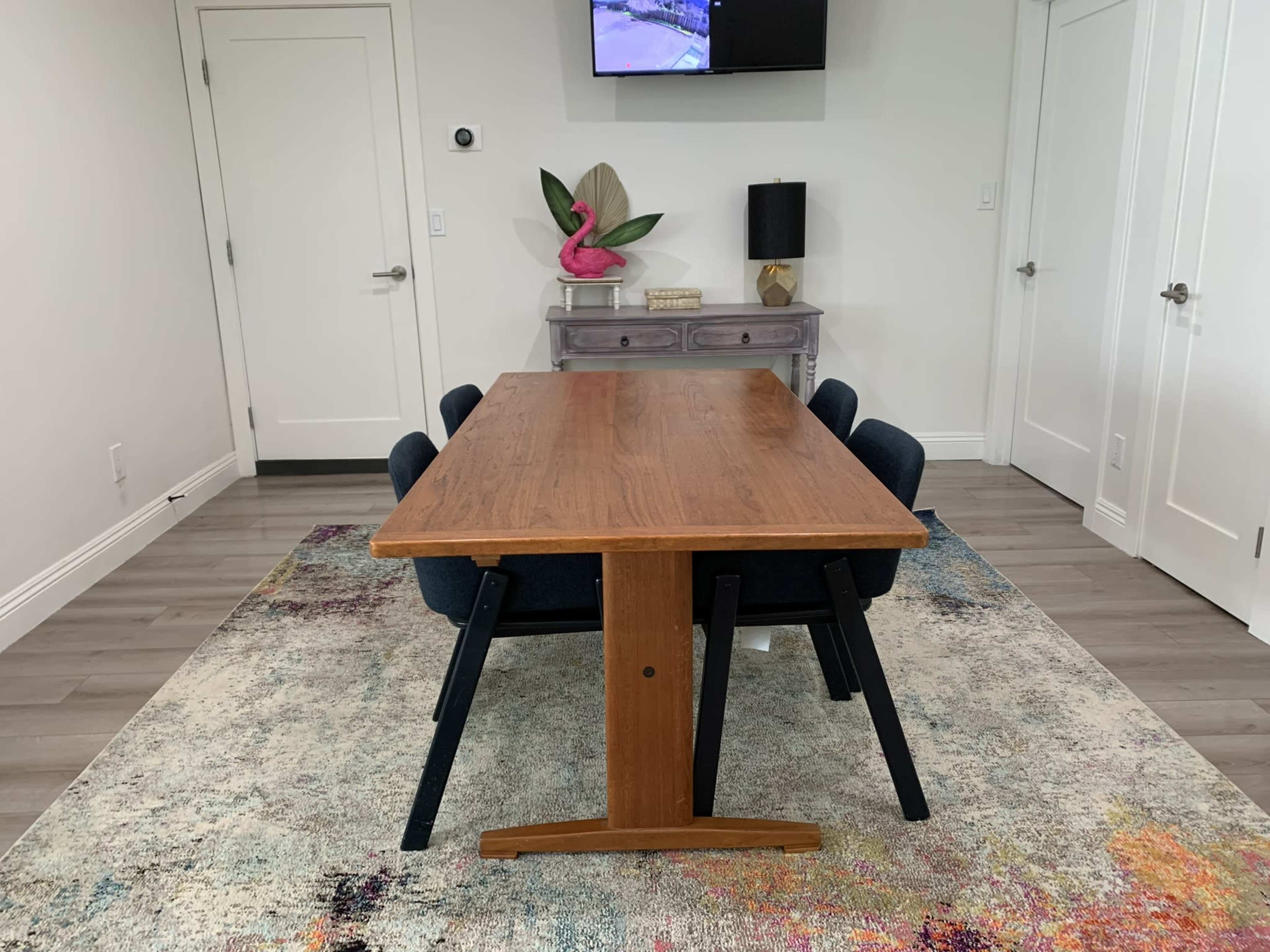 A wooden dining table is set in a room with blue chairs, a colorful rug, and a television mounted on the wall.