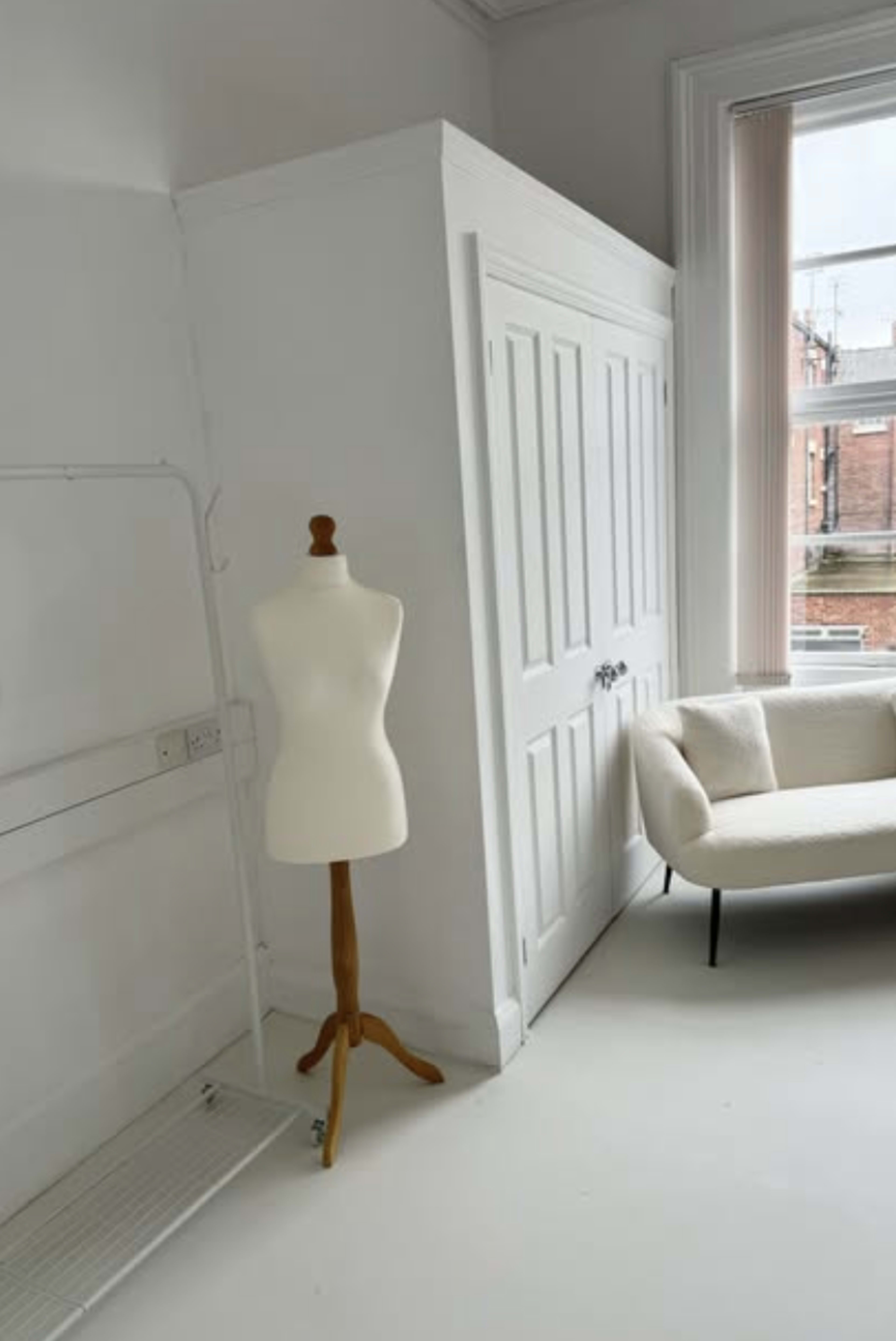 The image shows a minimalist room with a white mannequin on a stand, a simple couch, and white walls and cabinetry.
