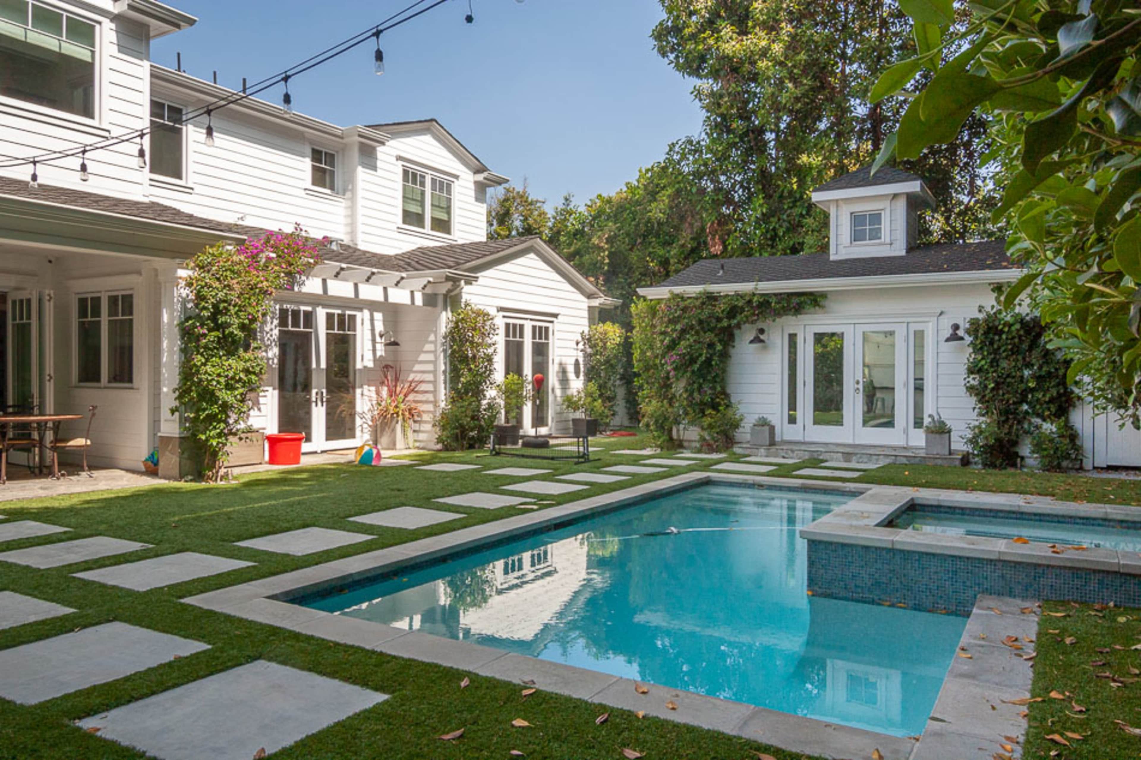 A modern backyard with a swimming pool surrounded by landscaped greenery and a white house in the background.