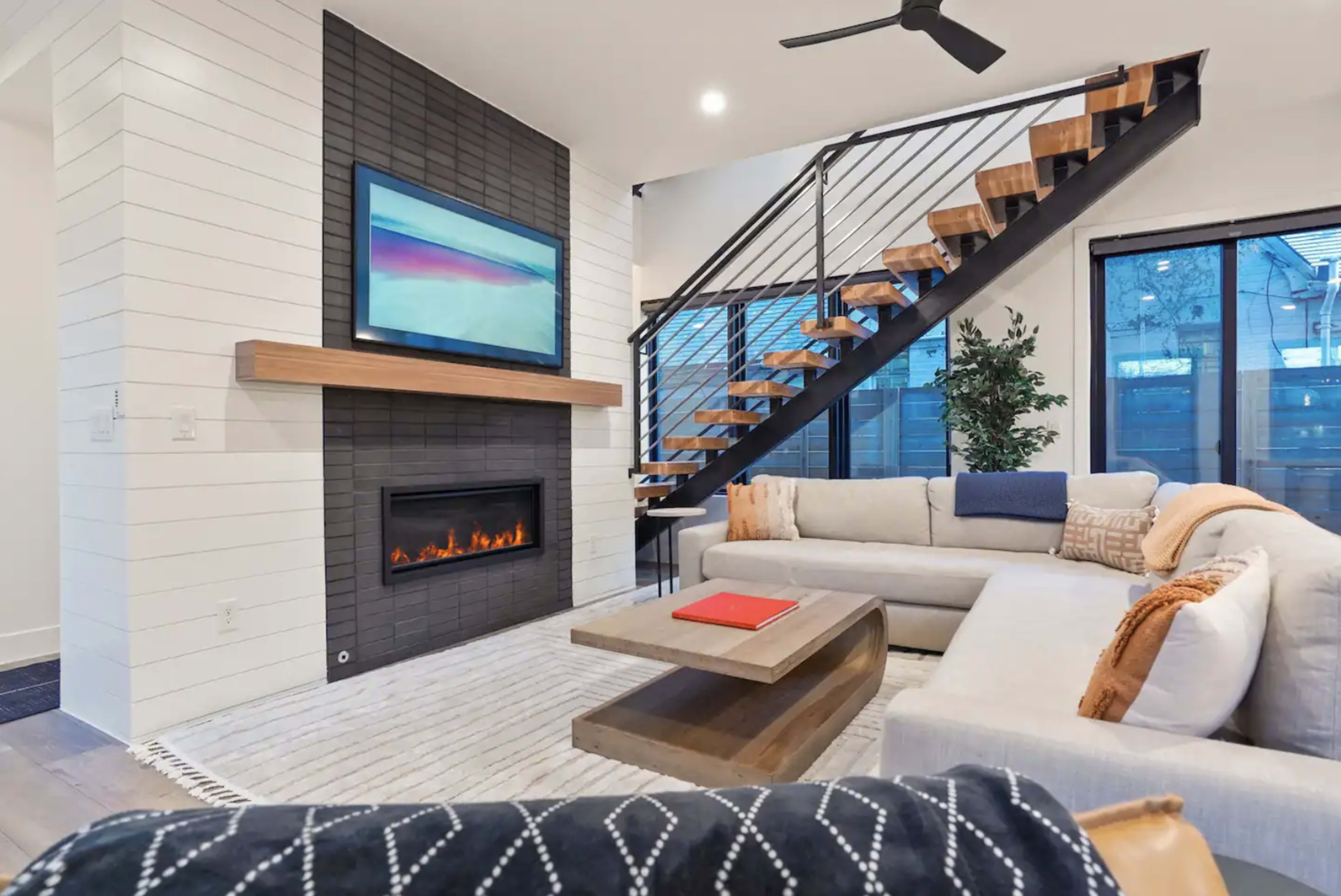 A modern living room with a sectional sofa, a coffee table, a wall-mounted television above a fireplace, and a staircase leading to an upper floor.