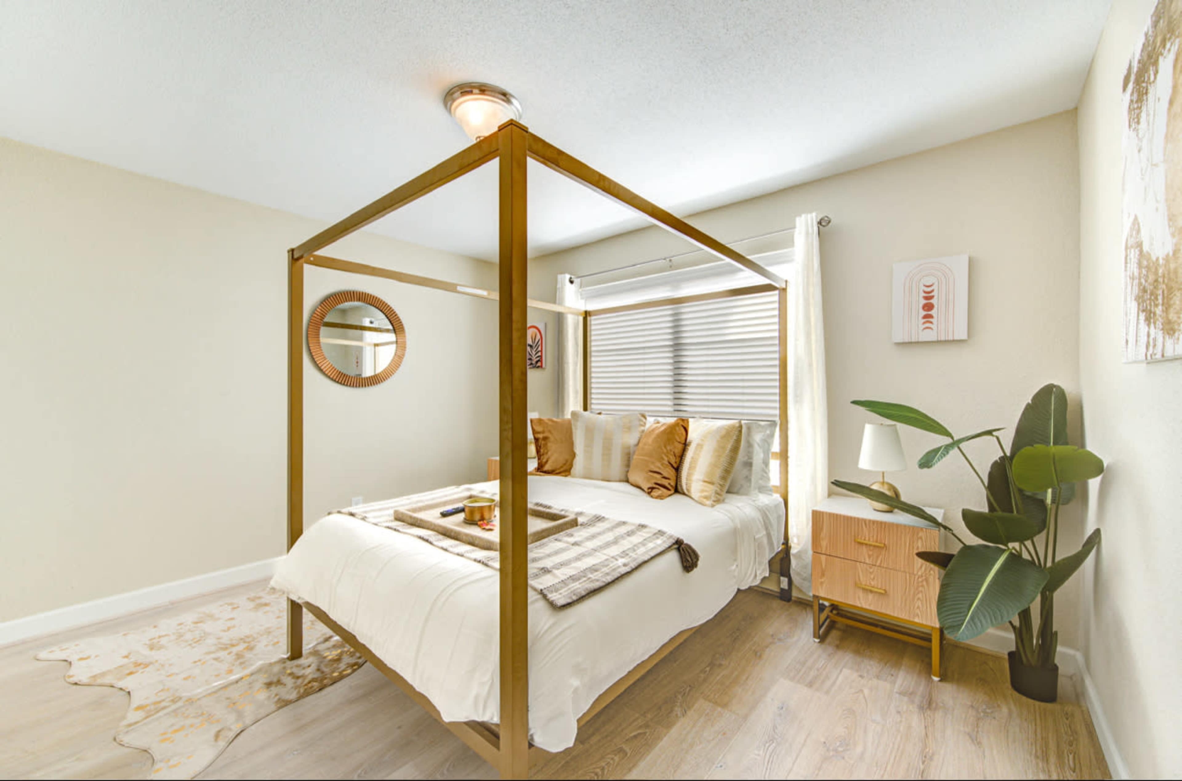 The bedroom features a four-poster bed with a light-colored comforter, a nightstand, and a decorative plant in a softly lit space.