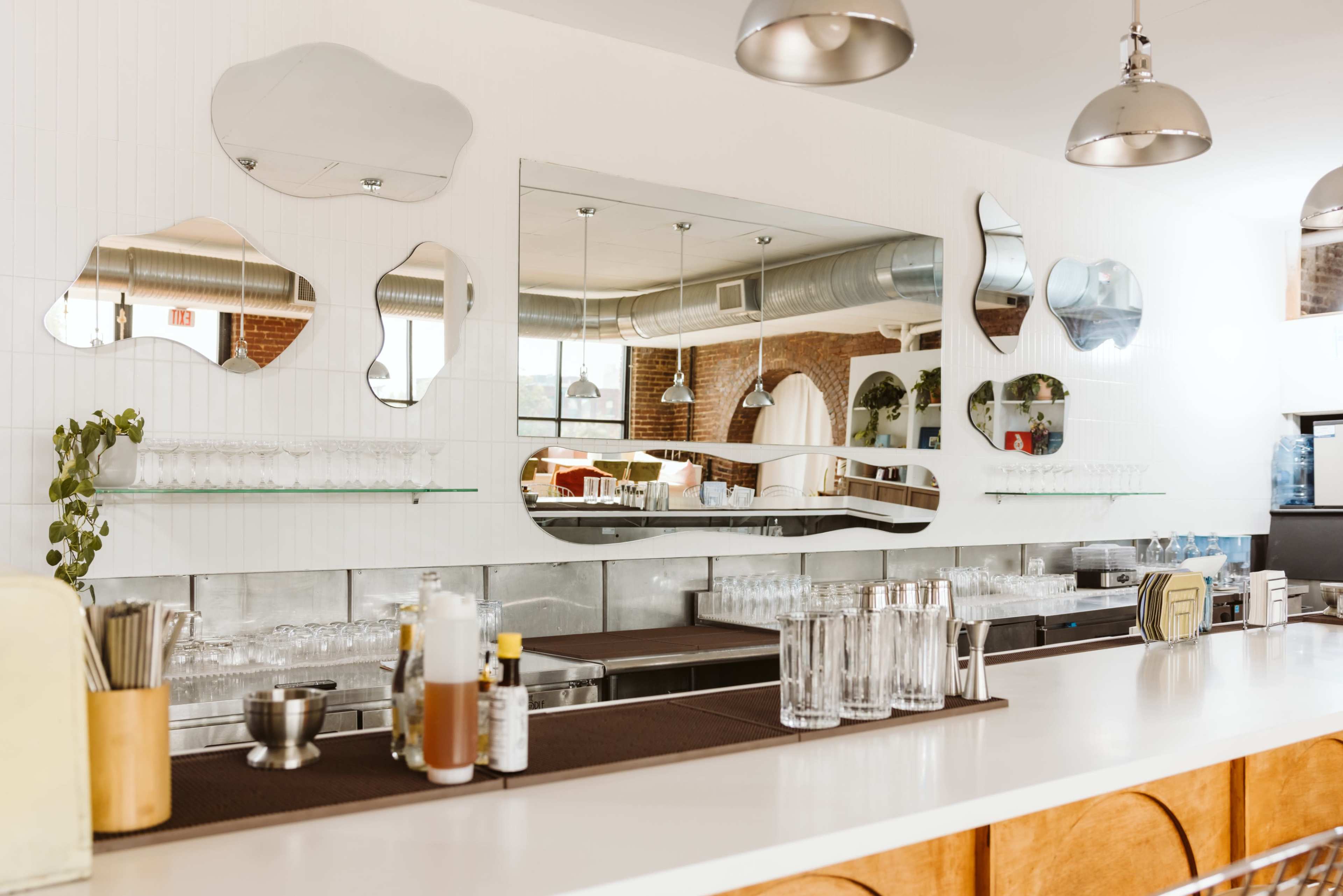 The image shows a modern bar area featuring a long counter, mirrors with organic shapes on the wall, and shelves displaying glassware and drink ingredients.