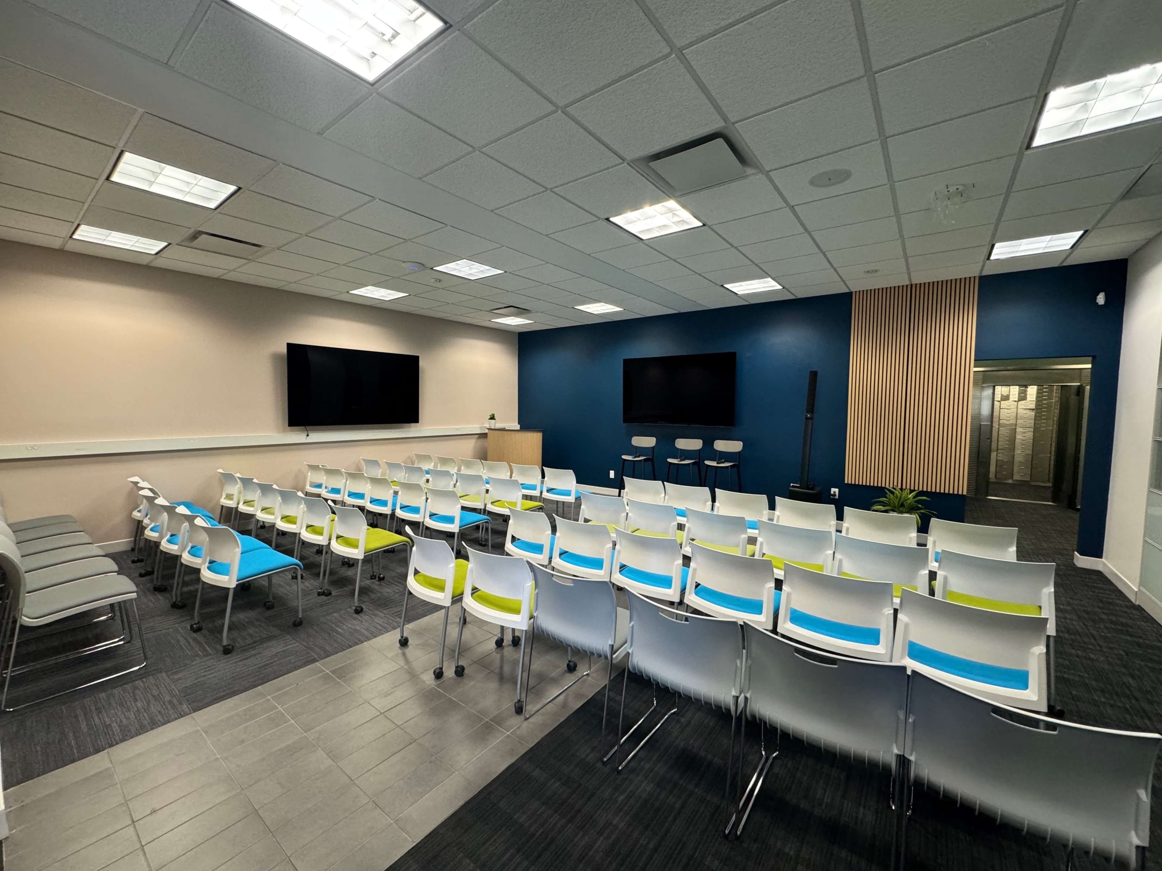 A modern conference room with rows of white chairs featuring colored accents, two large screens on the walls, and a small stage area.