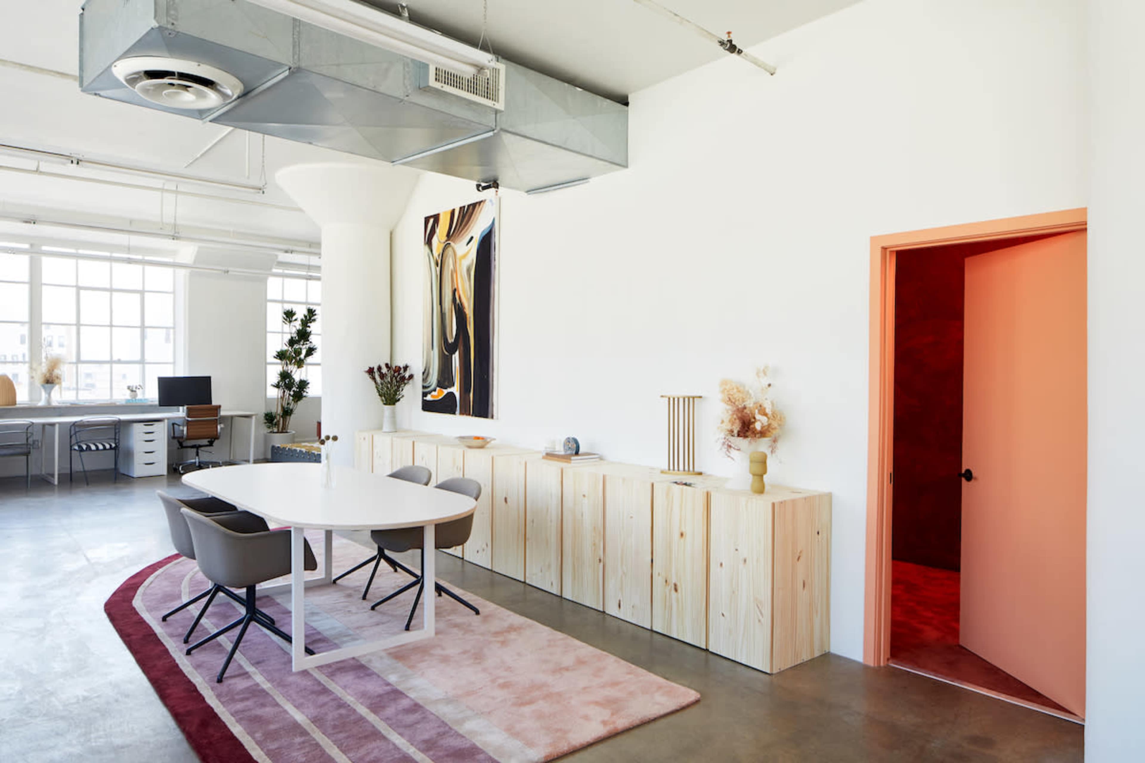 A modern office space features a white dining table with gray chairs, wooden cabinetry, and an orange door leading to another room.