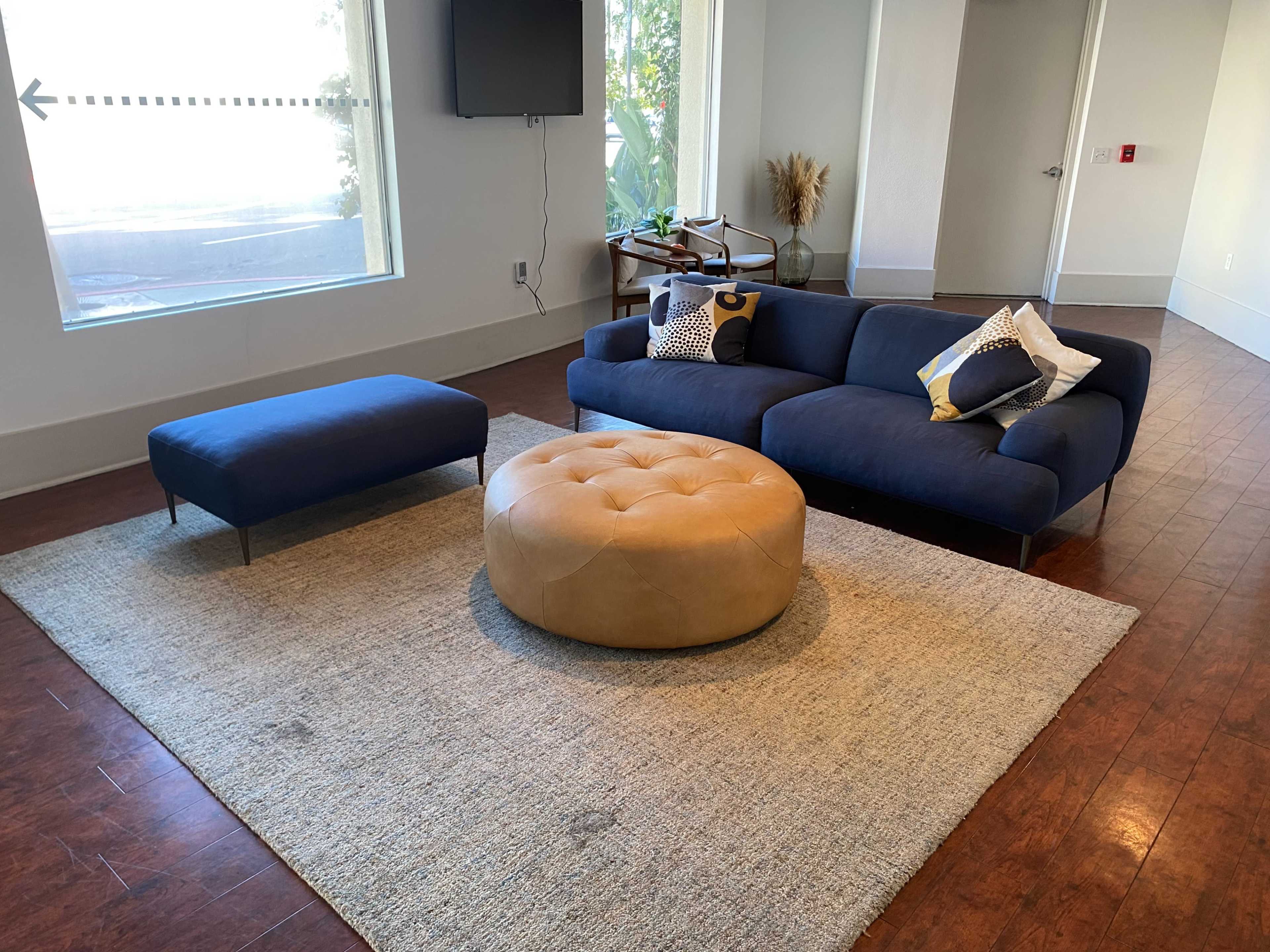 The image shows a modern living room with a blue sectional sofa, a round tan ottoman, and a large area rug, all arranged near a window.