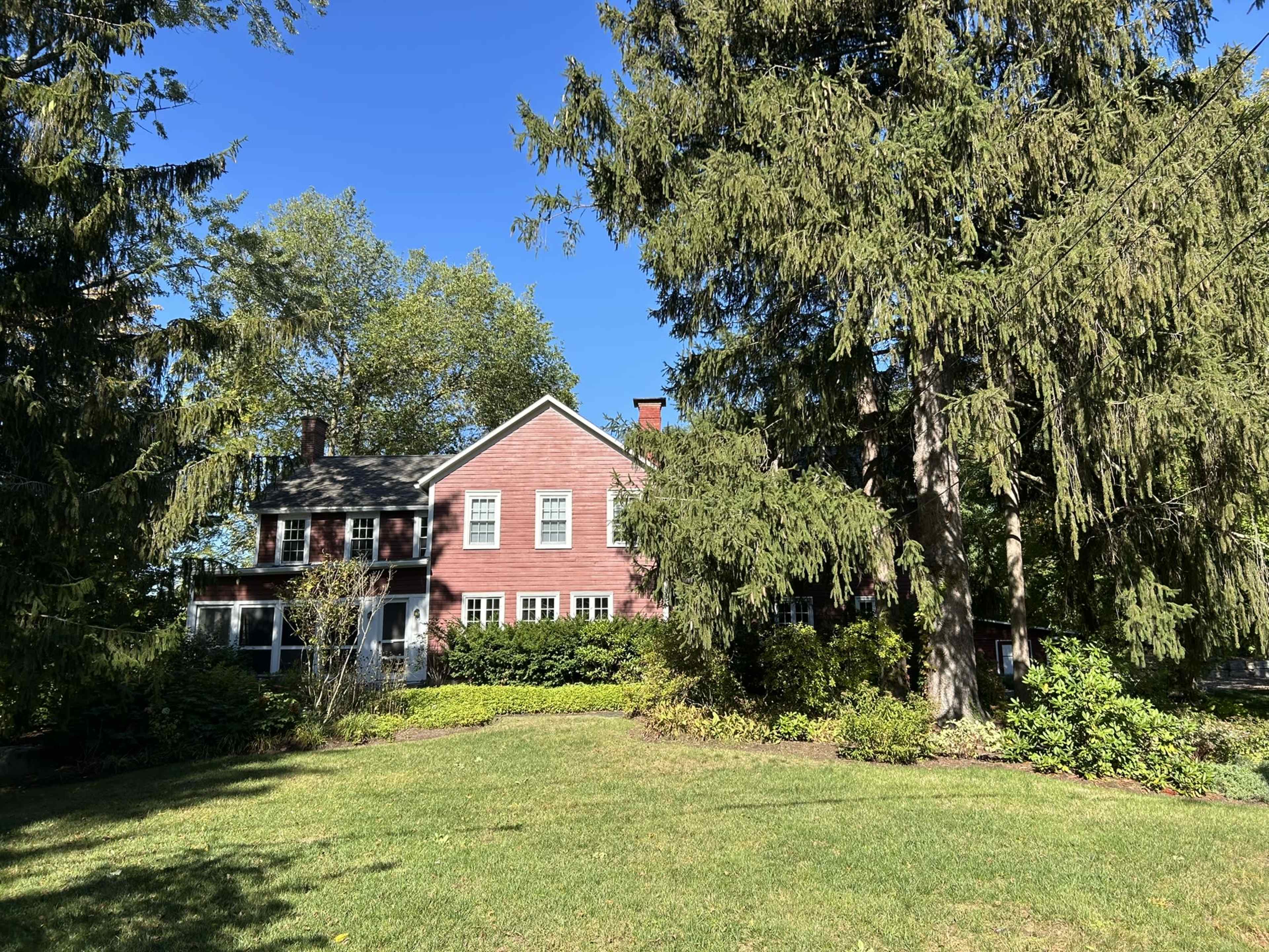 A red house with white windows is surrounded by tall trees and a green lawn.