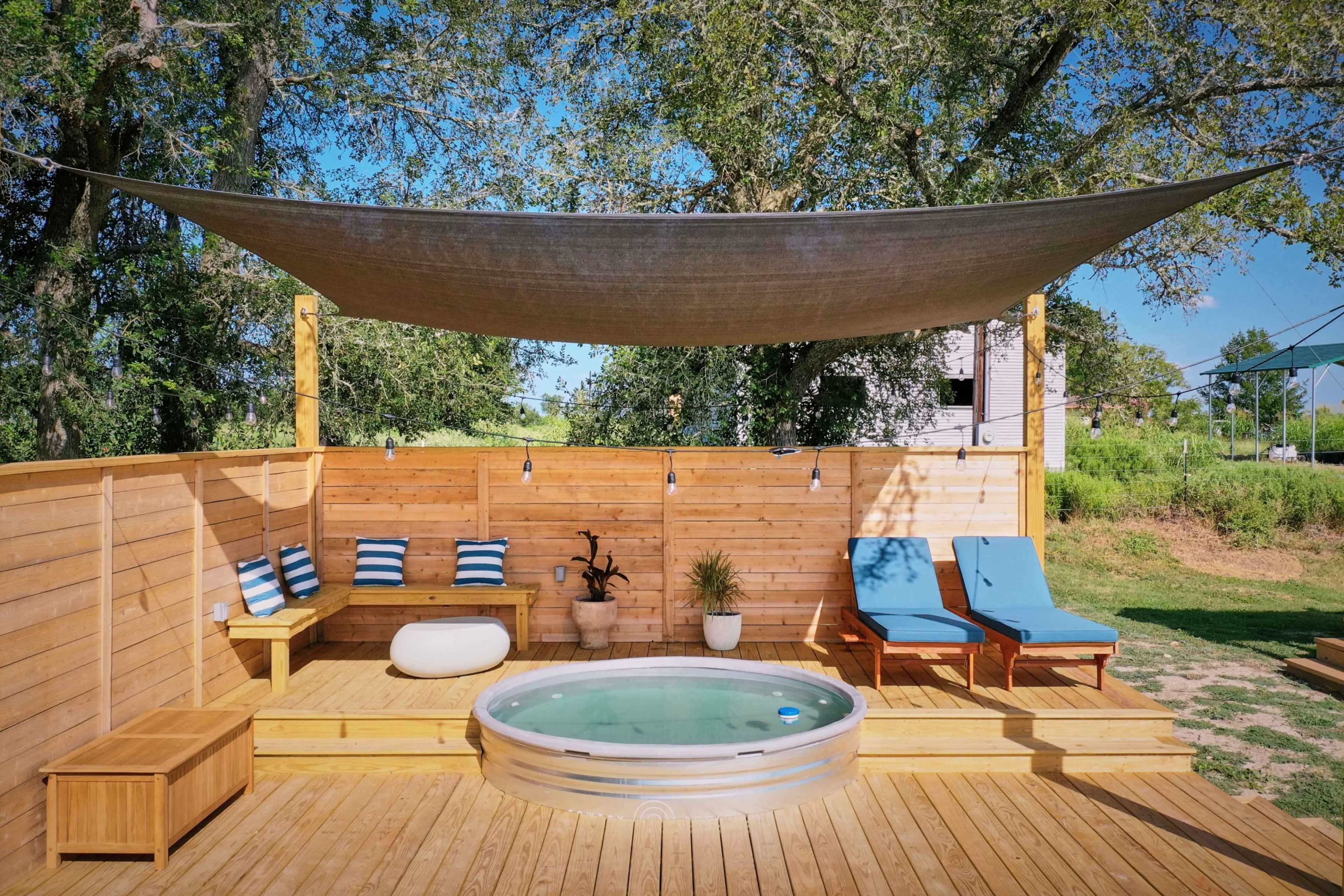 The image shows a wooden deck with a hot tub, two blue lounge chairs, seating with cushions, and a shaded area under a canopy.