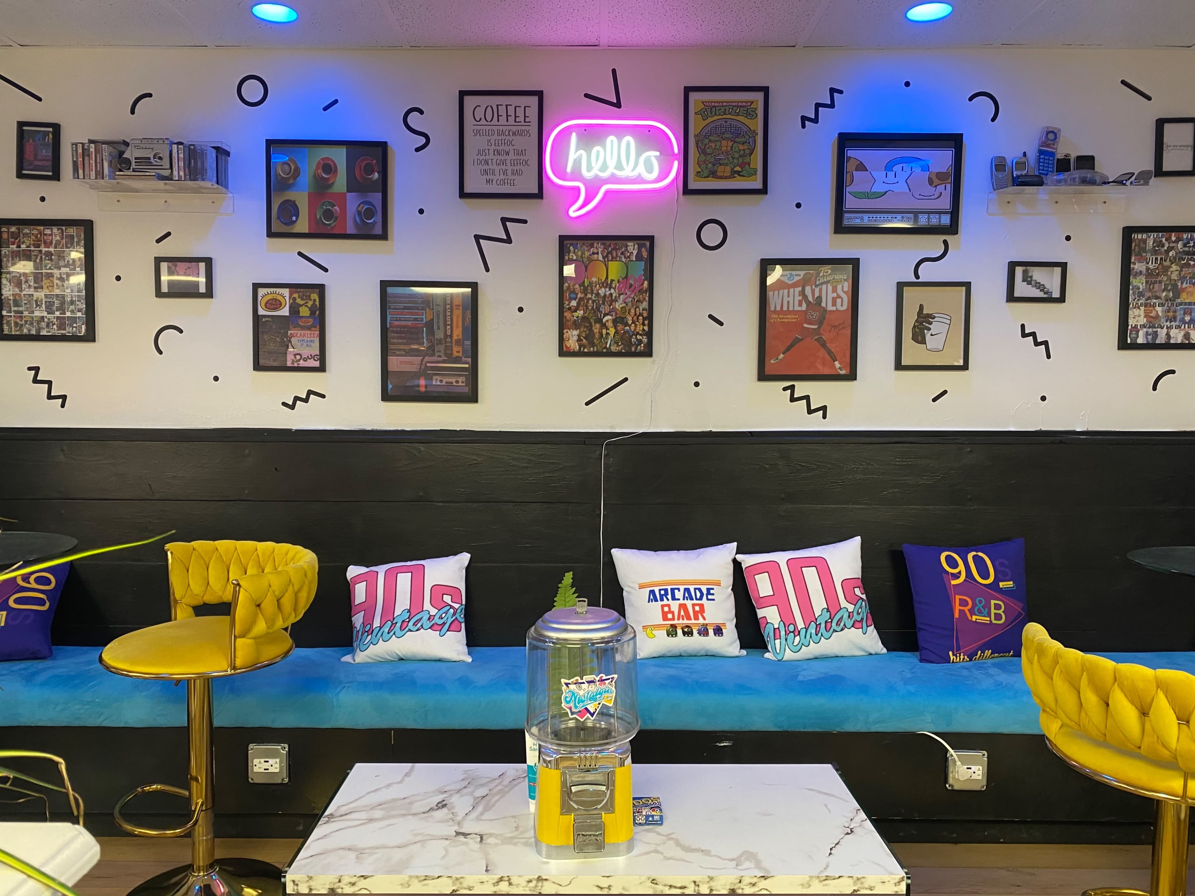 A colorful café interior features yellow chairs, a blue banquette, and walls adorned with framed artwork and neon signage.