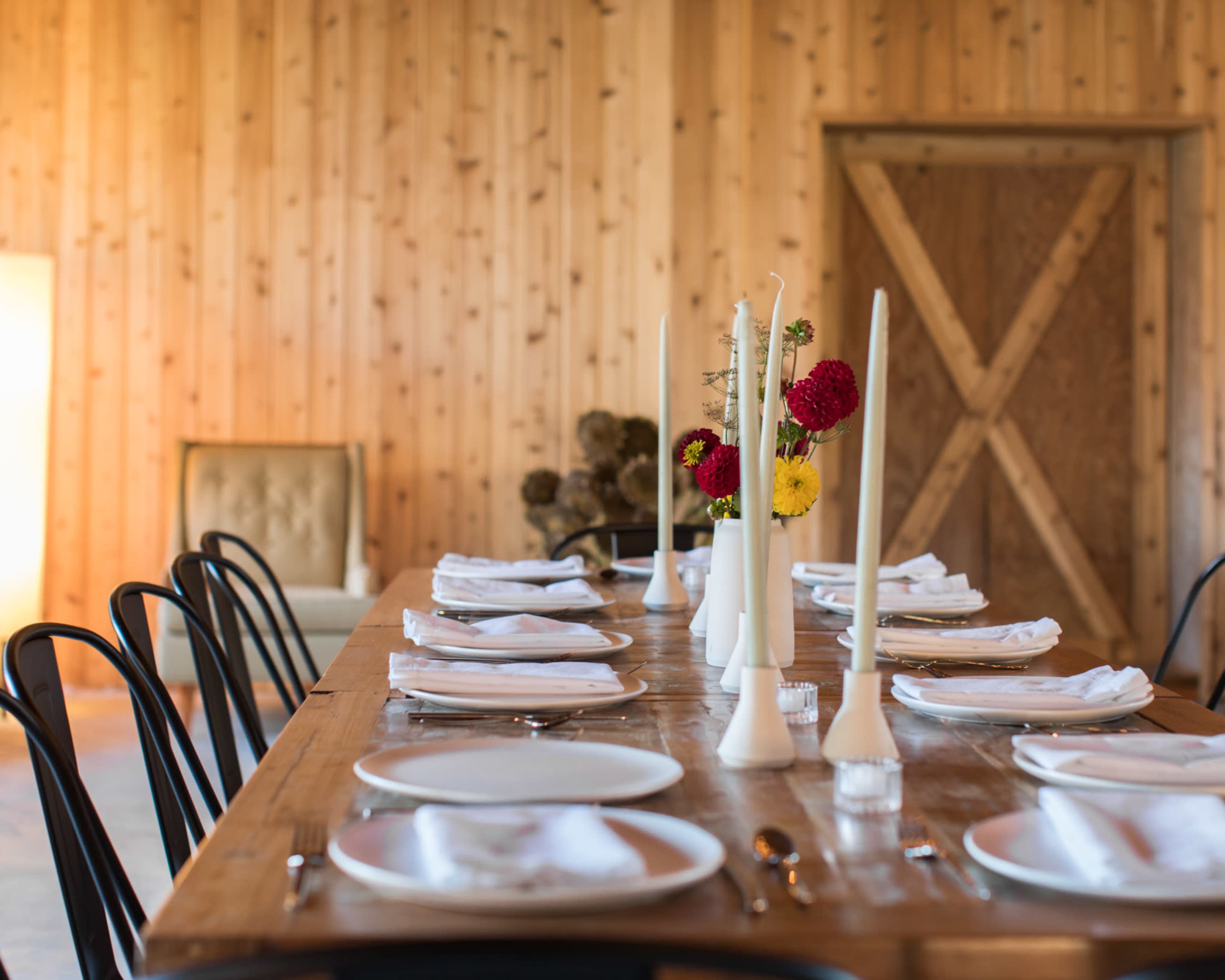 A long wooden dining table is set with white plates and candles, complemented by a bouquet of flowers in the center and surrounded by black chairs.