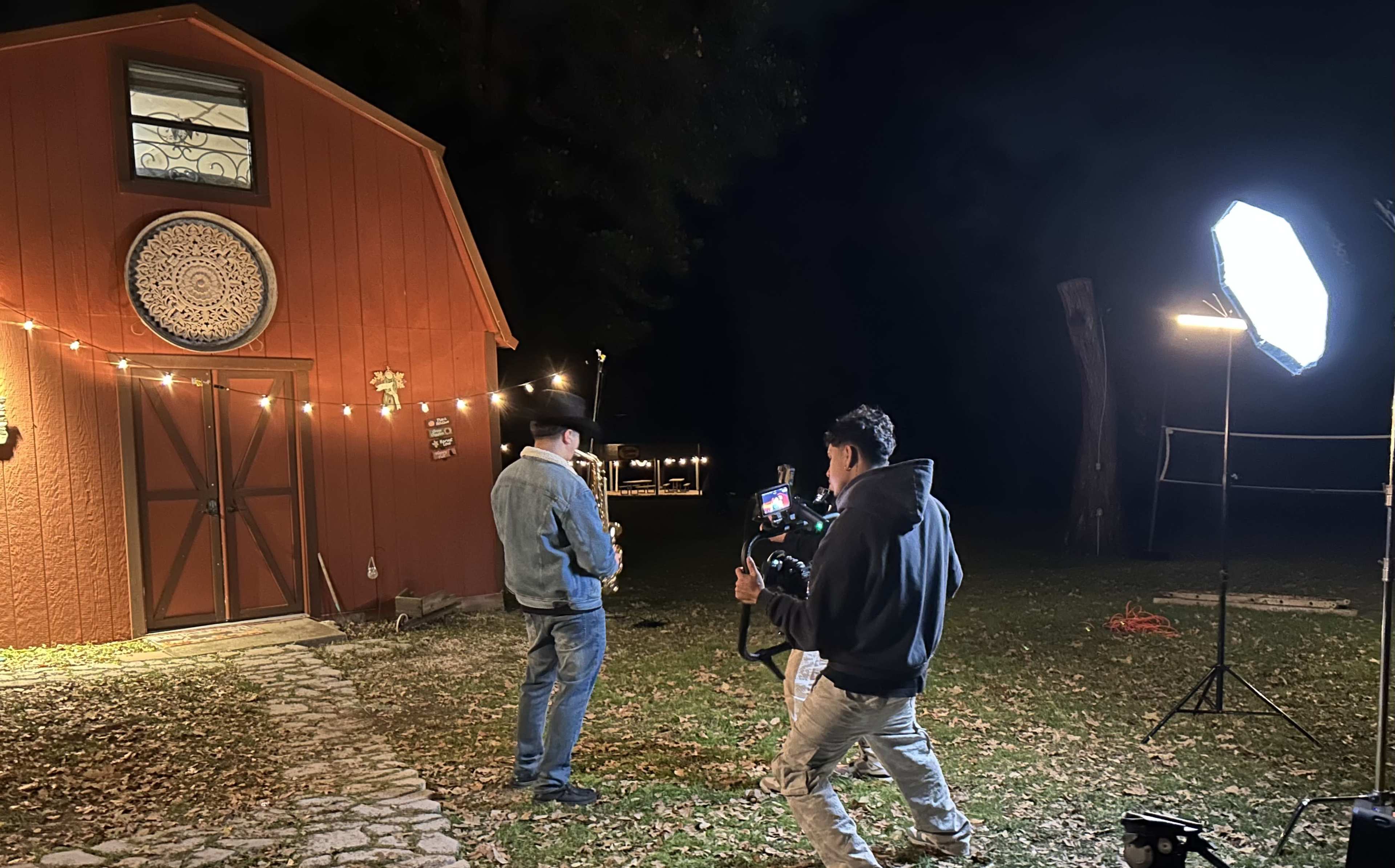 A man in a cowboy hat is playing guitar outside a barn during night filming, while a second man operates a camera with lights set up nearby.