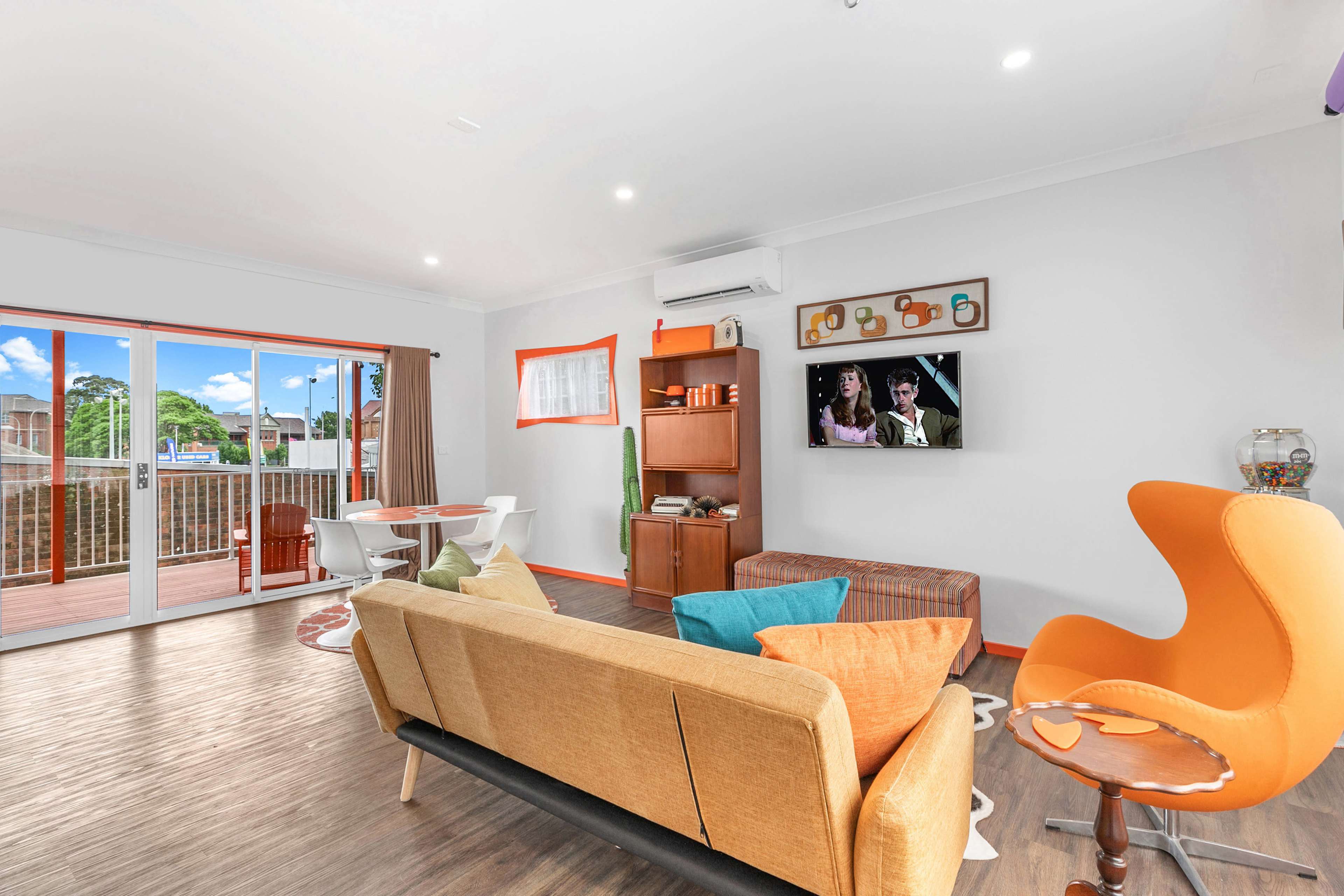 A modern living room features a sofa, an orange armchair, a television on the wall, and a dining area with a round table and chairs.