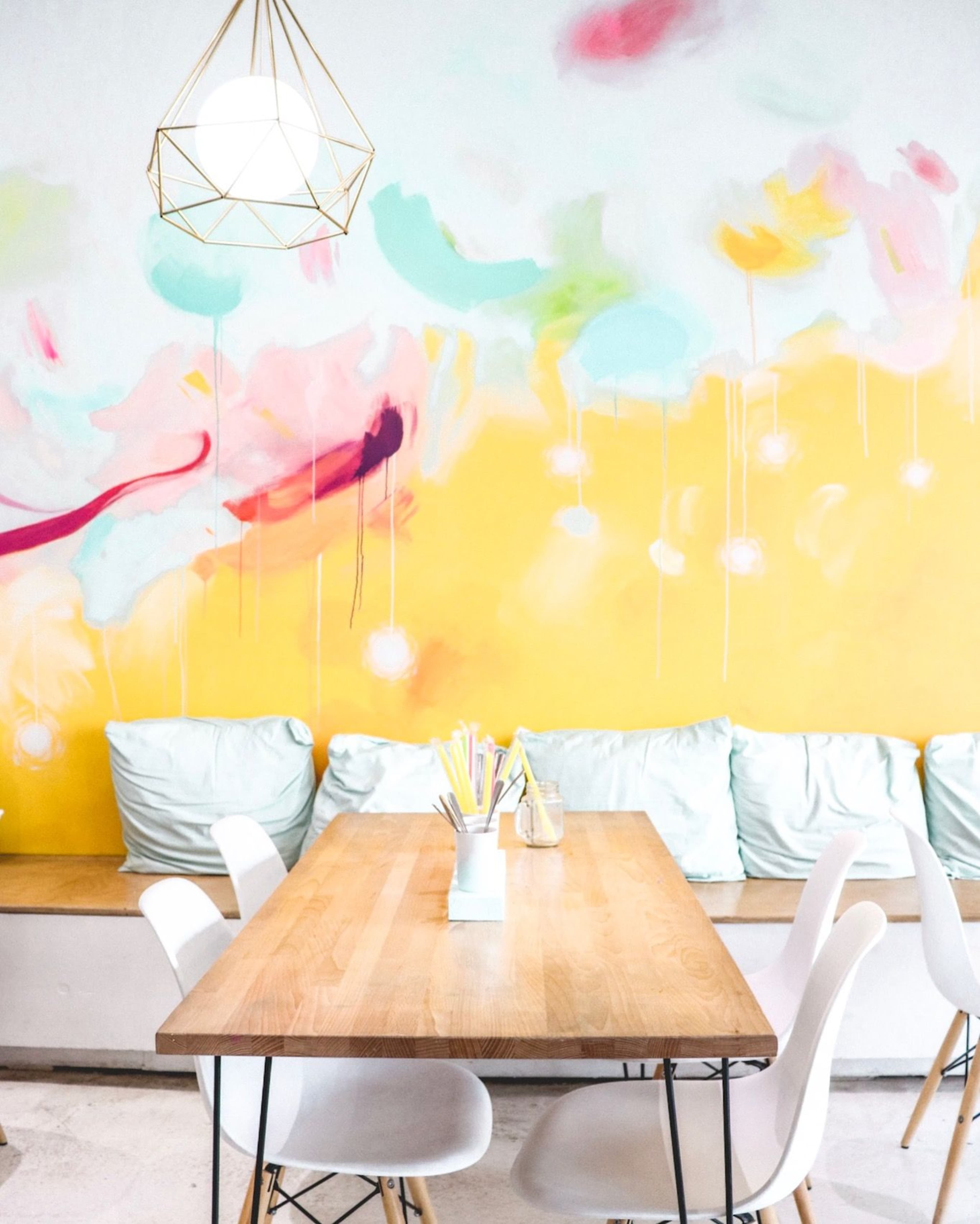 A wooden table sits in front of a brightly colored mural, with white chairs and a seating area featuring light blue cushions.