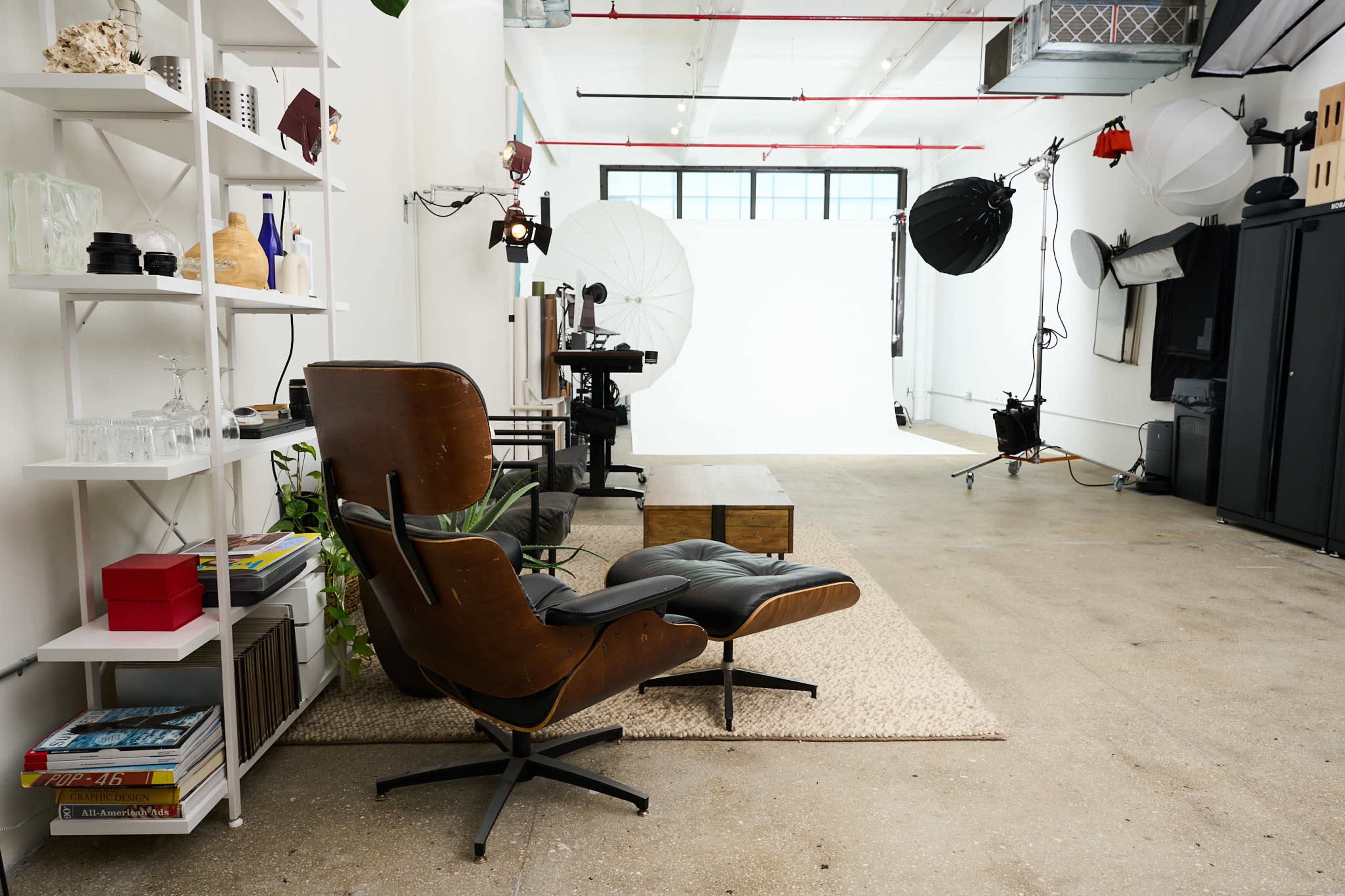 The image shows a spacious studio setup with a leather chair, a wooden coffee table, and various photography equipment arranged against a white backdrop.
