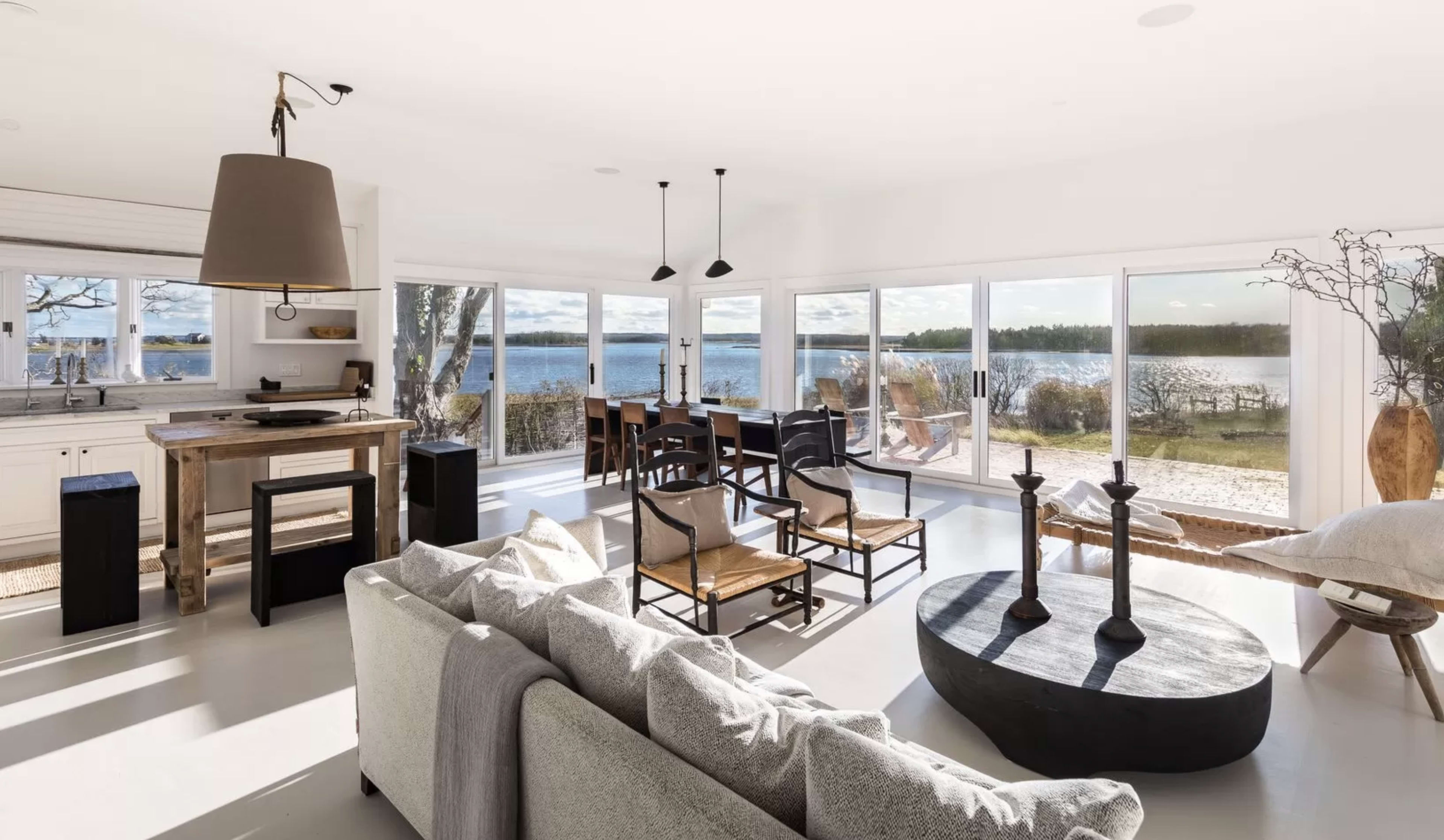 A modern open-concept living area features a spacious layout with large windows overlooking a serene waterfront, furnished with a gray sofa, wooden dining set, and decorative elements.