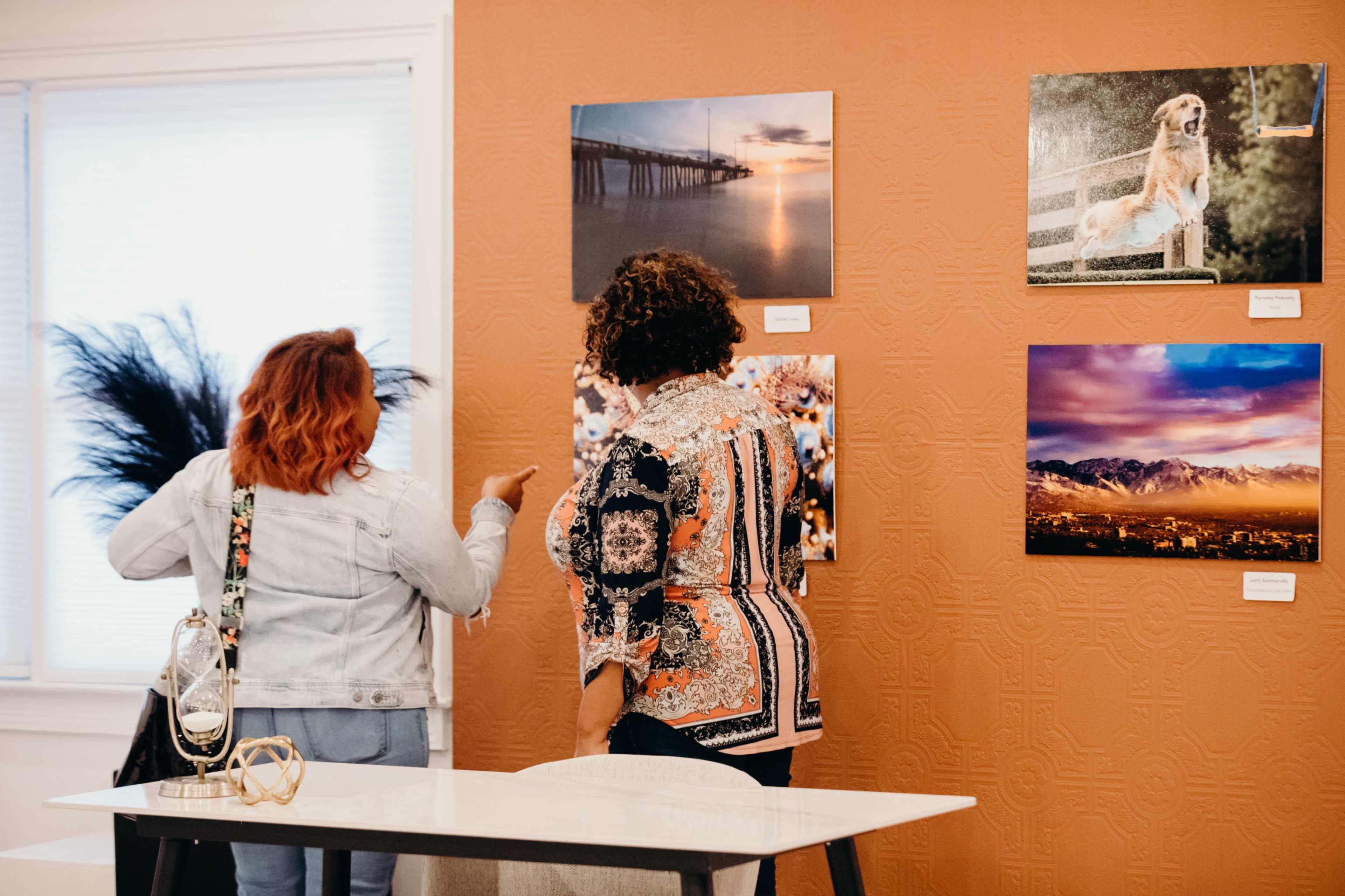 Two women observe photographs displayed on an orange wall in an art gallery setting.